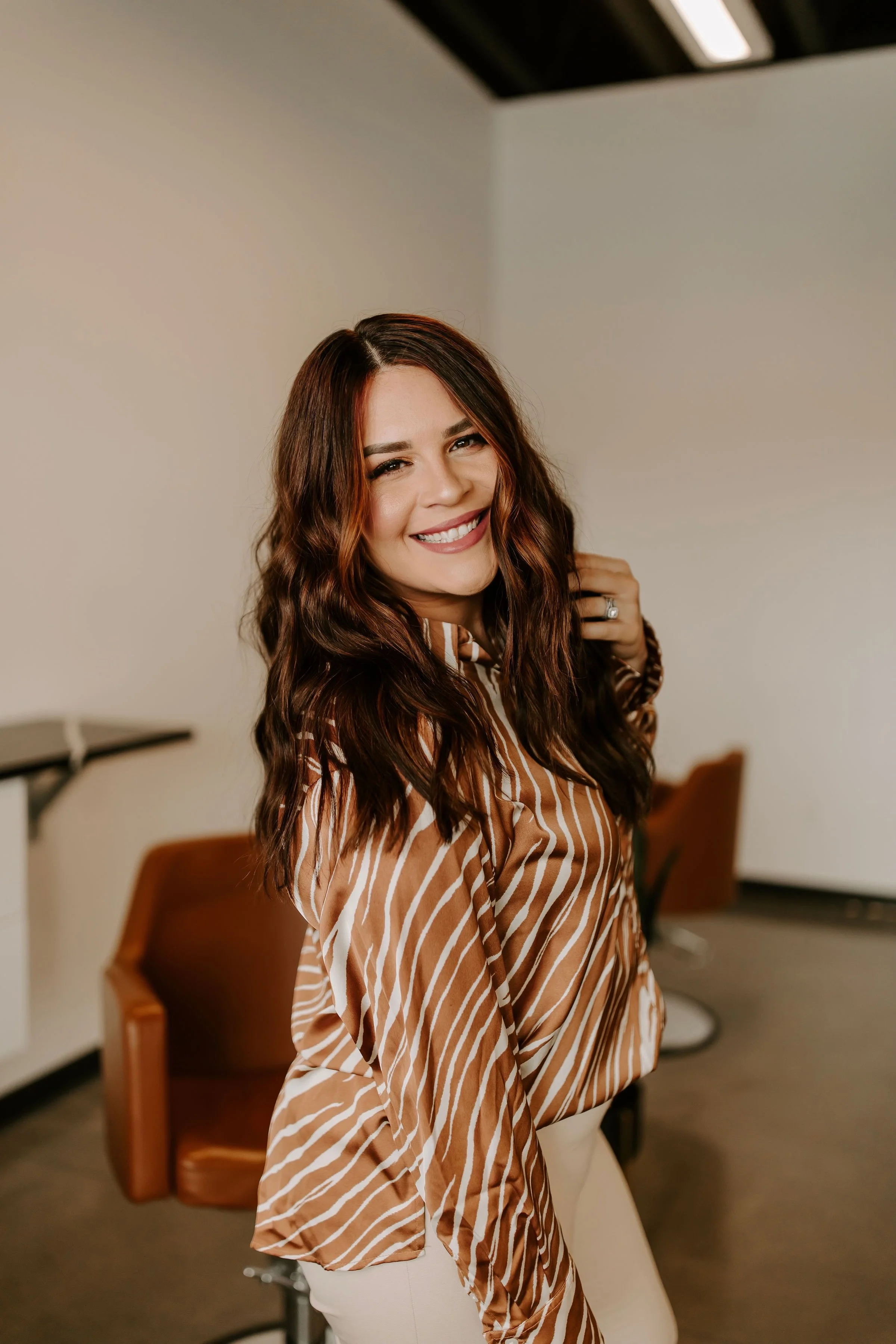 A young woman with long, wavy, reddish-brown hair smiling and looking at the camera, standing in an indoor space with chairs and a white wall in the background.