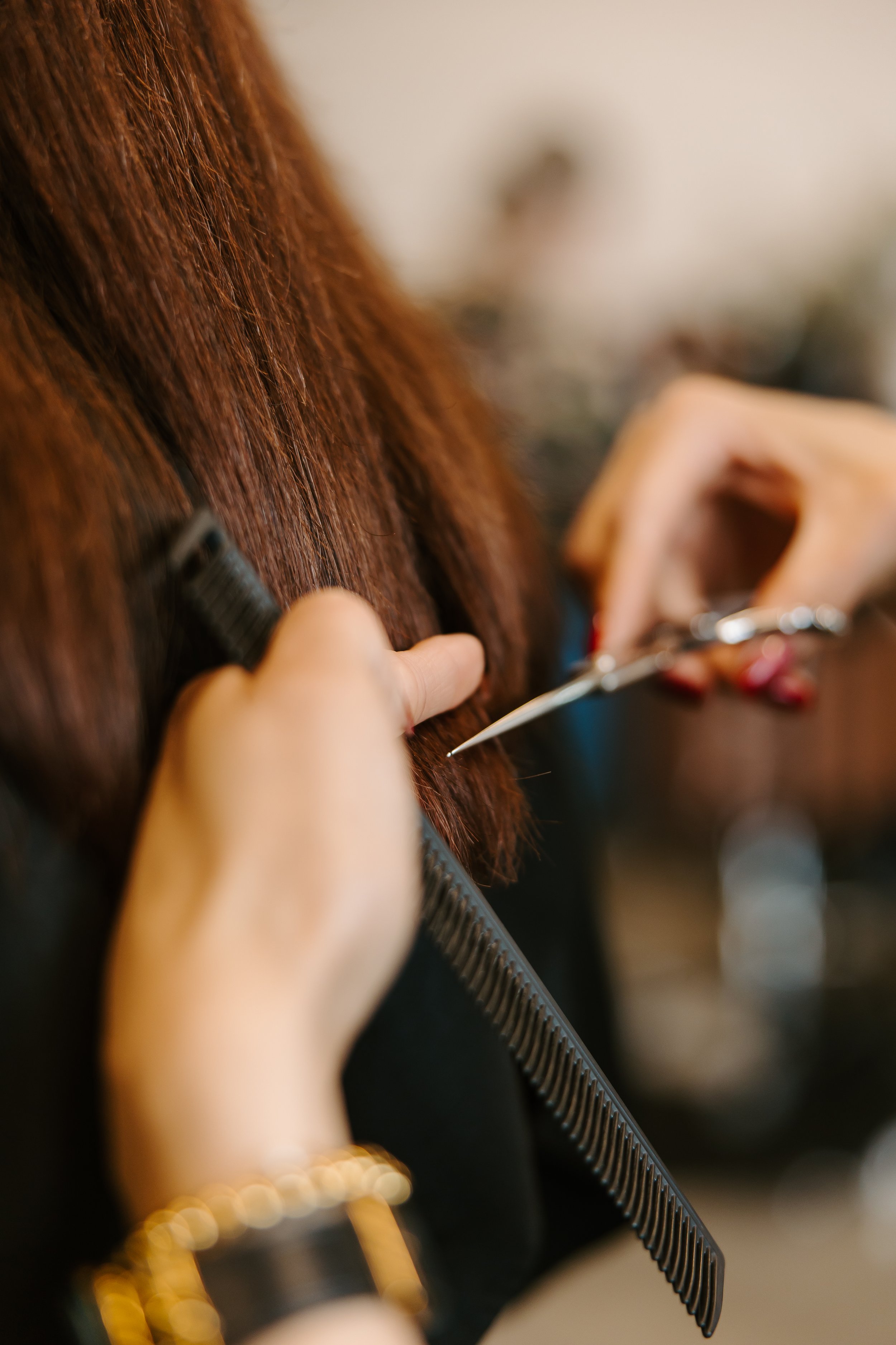 A hairstylist is cutting a woman's reddish-brown hair with scissors, holding a comb in their other hand.