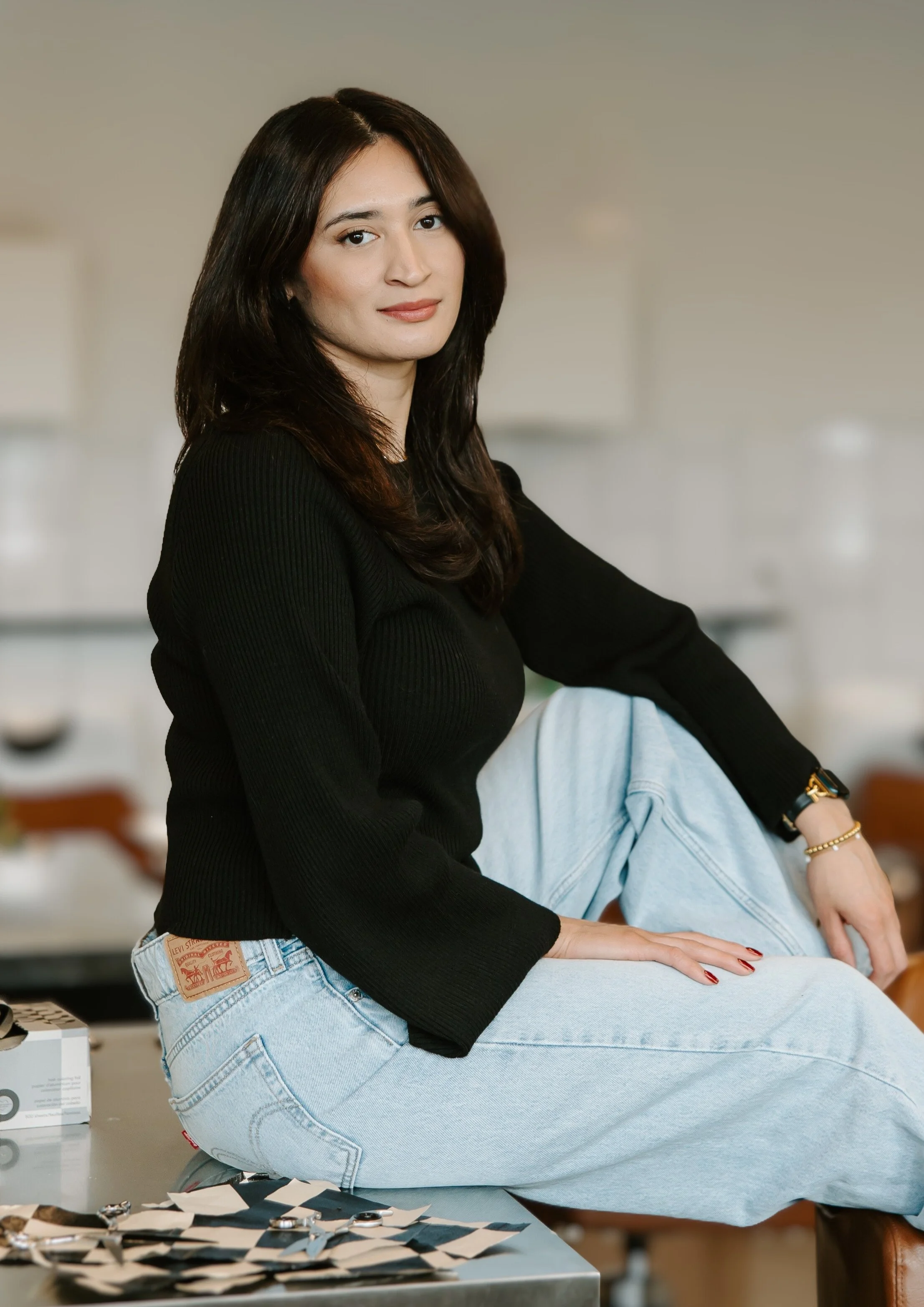A woman with dark brown hair wearing a black top and light blue jeans, sitting on a table in a bright room.