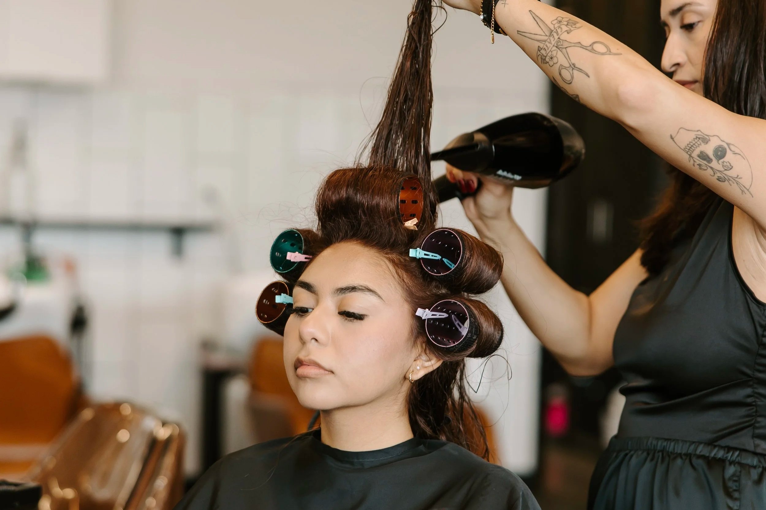 A woman with hair rollers at a salon getting her hair blow-dried by a stylist.