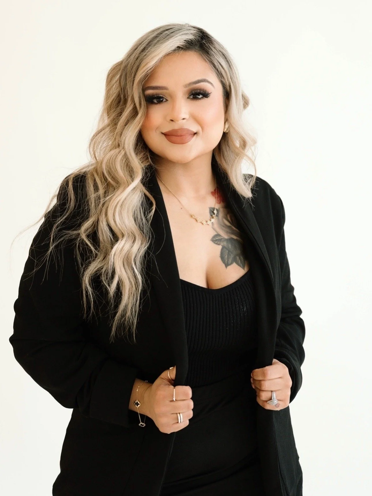 A woman with long, wavy blonde hair, wearing makeup, a black top, and a black blazer, standing against a plain light background.