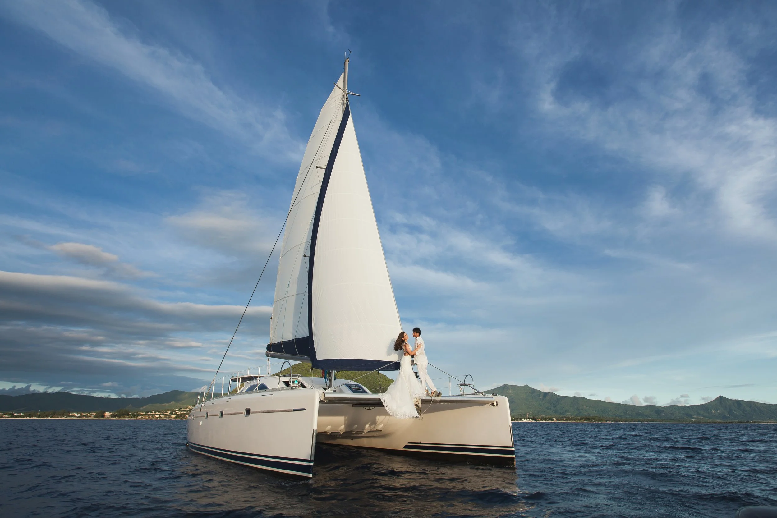 A couple dressed in wedding attire standing on a sailing yacht with a large sail, on the water with a mountain range in the background under a partly cloudy sky.
