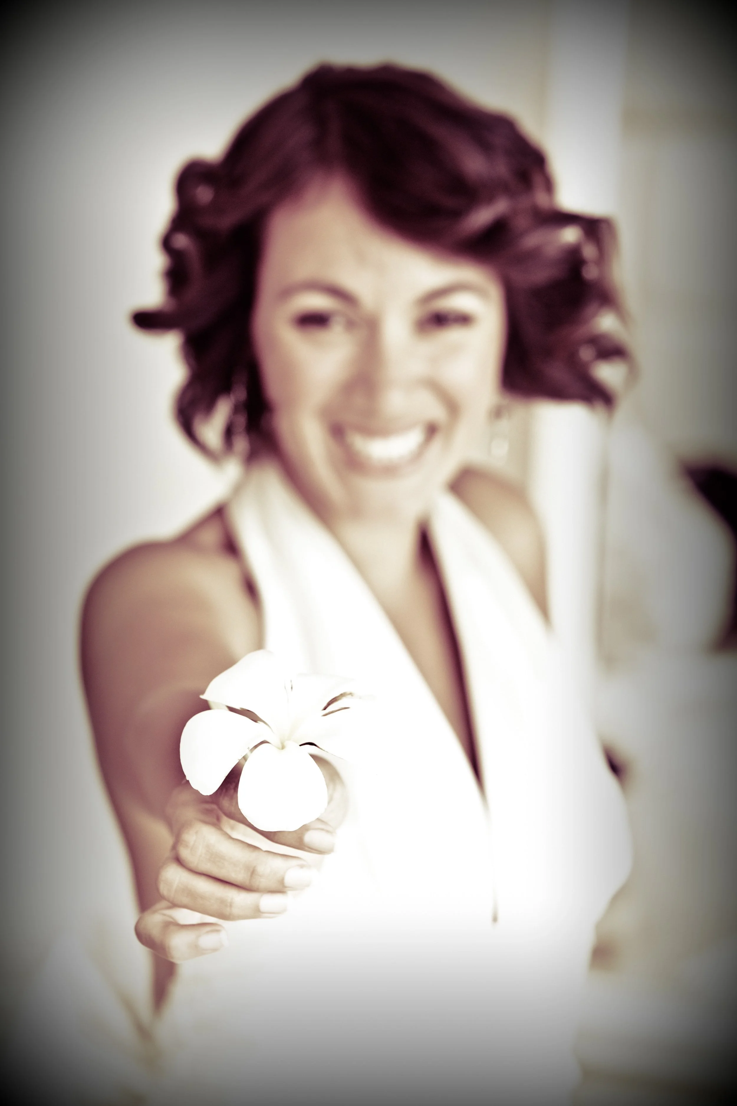 A woman with curly hair smiling and holding a white flower, extending her arm toward the camera.