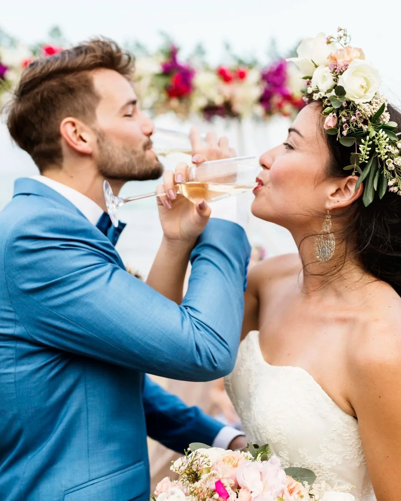 A groom and bride sharing a toast at their wedding, with the groom in a blue suit and the bride wearing a white dress and floral headpiece, holding a bouquet, outdoors with colorful flowers in the background.