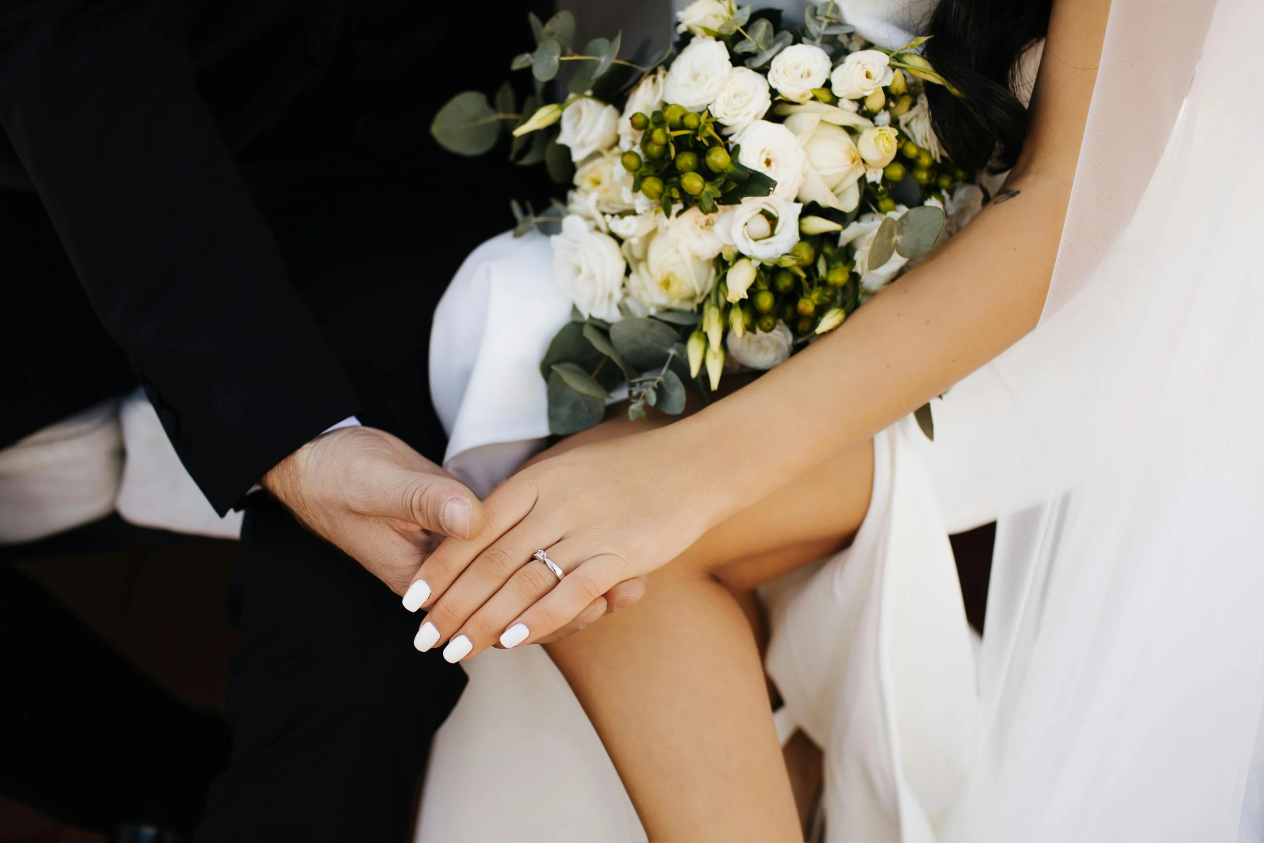 Close-up of a bride and groom holding hands, with a bouquet of white flowers and greenery, wedding ring visible on the bride's finger.