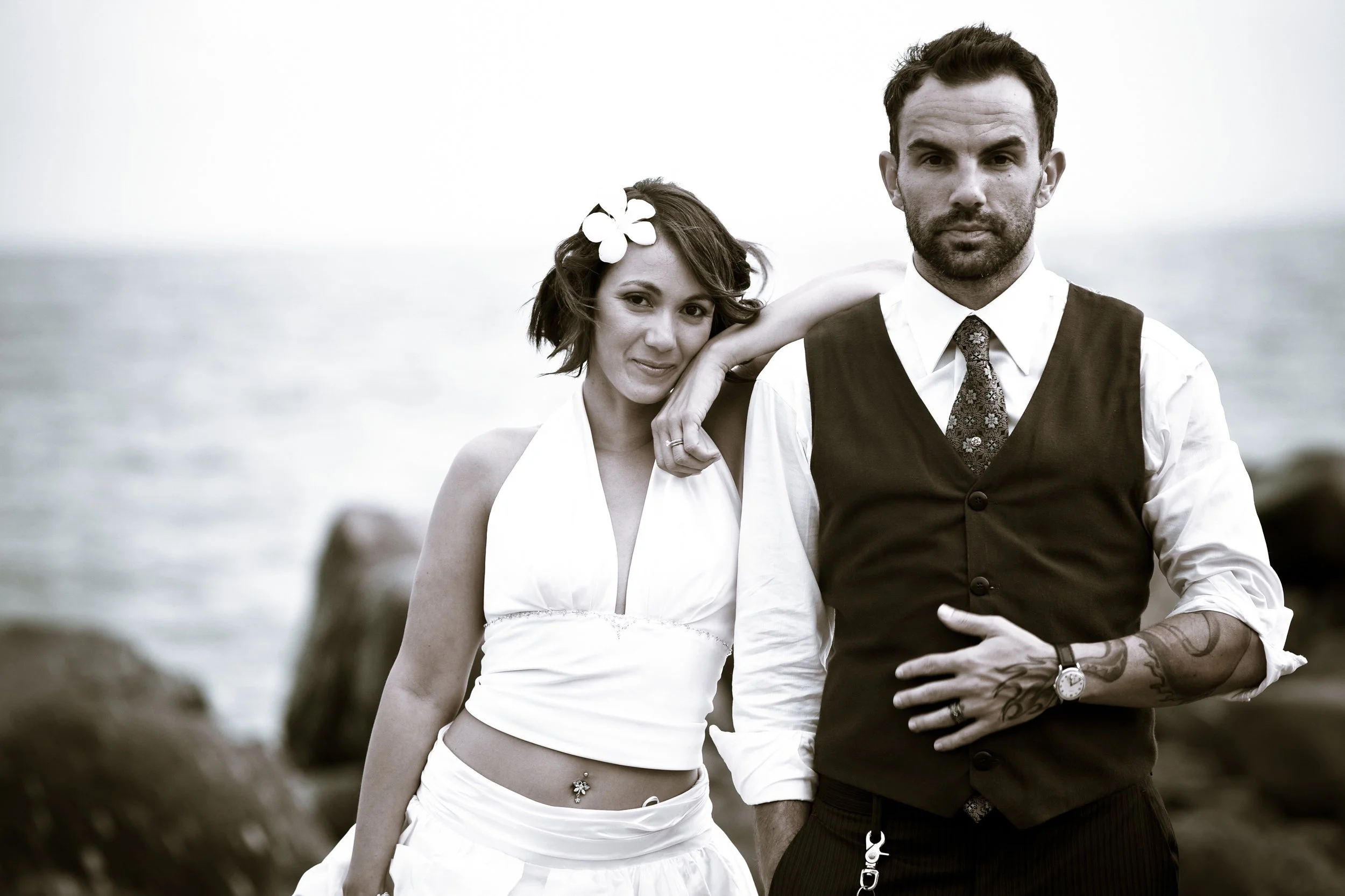 A black and white photo of a woman and man standing near the water, with rocky shoreline in the background. The woman has short hair, a floral hair accessory, a white sleeveless top, and a navel piercing. The man has short hair, a beard, tattoos on h