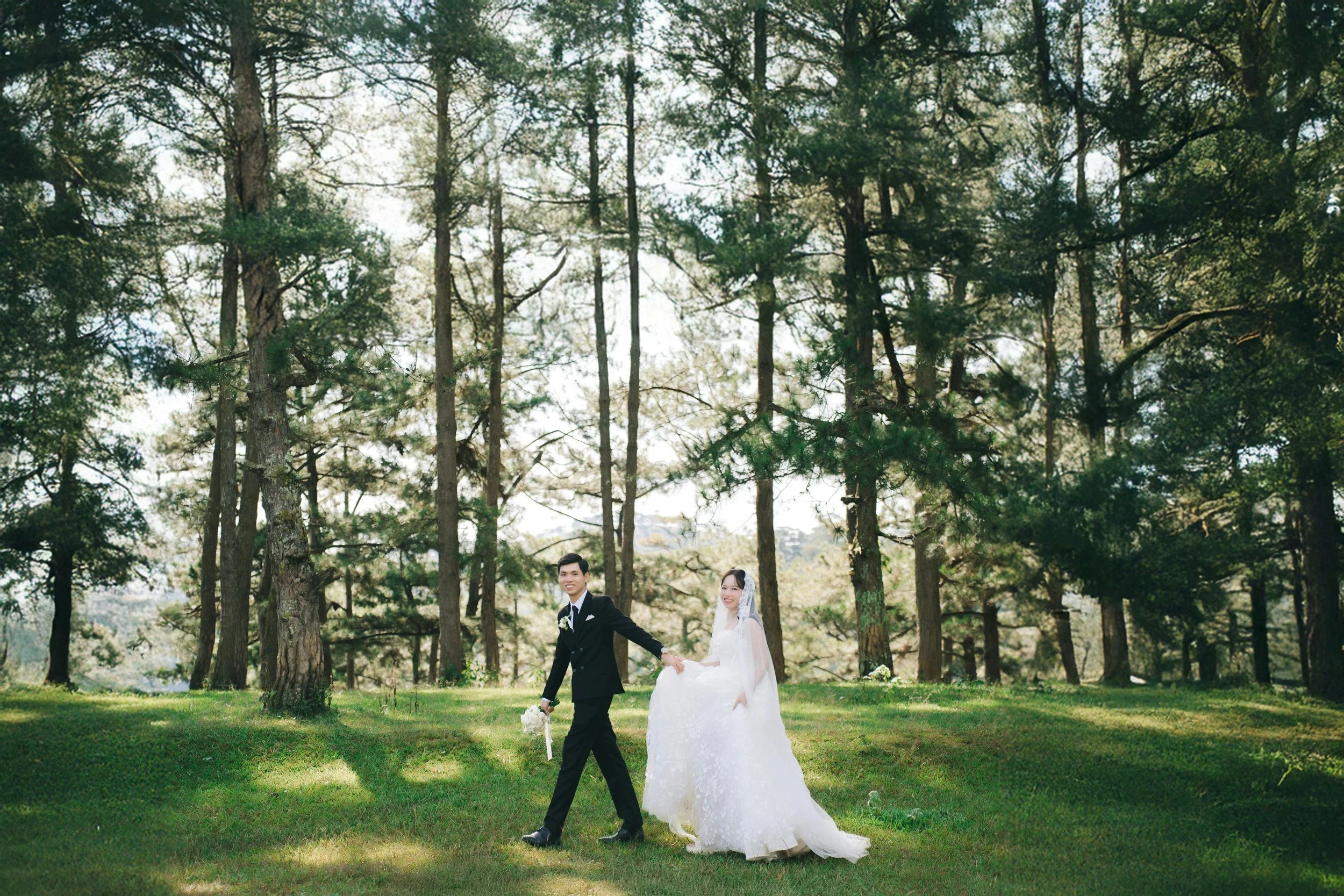 A bride and groom walking hand in hand in a forested area on their wedding day, with tall trees and green grass surrounding them.