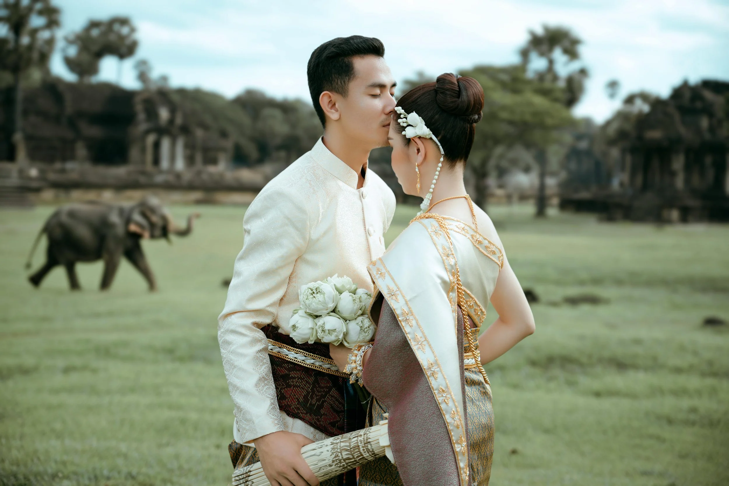 A newlywed couple in traditional costumes sharing an intimate moment outdoors with elephants in the background.