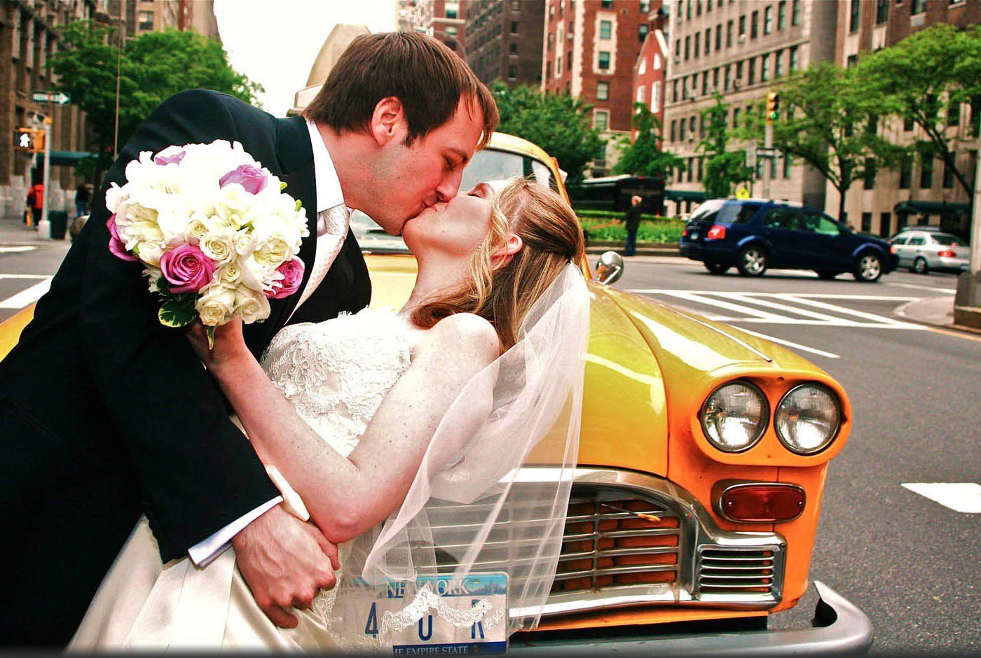 A bride and groom kissing on a city street in front of a vintage yellow taxi cab. The groom is holding a bouquet of pink and white flowers.