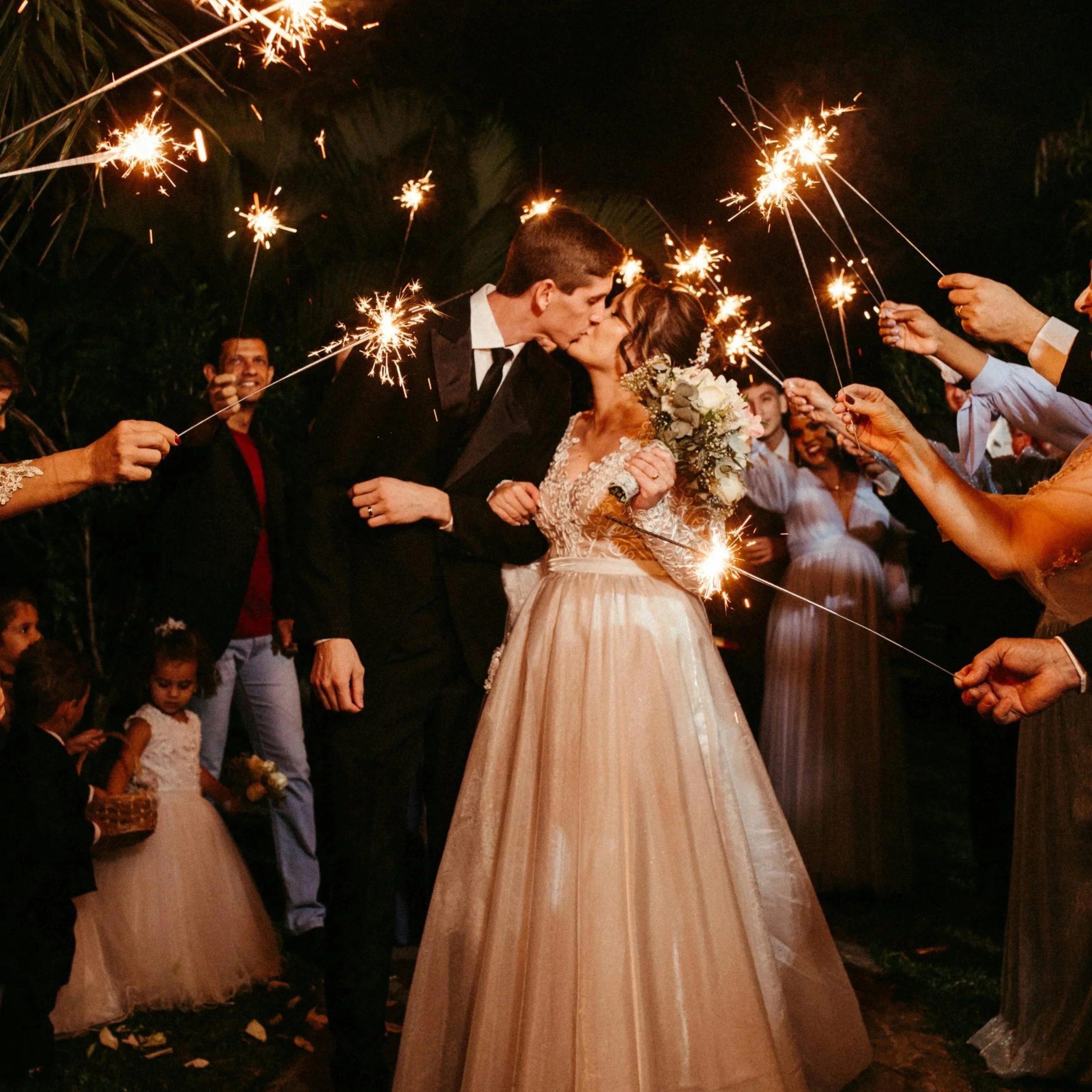 A newlywed couple shares a kiss under sparklers while surrounded by celebrating friends and family at night.