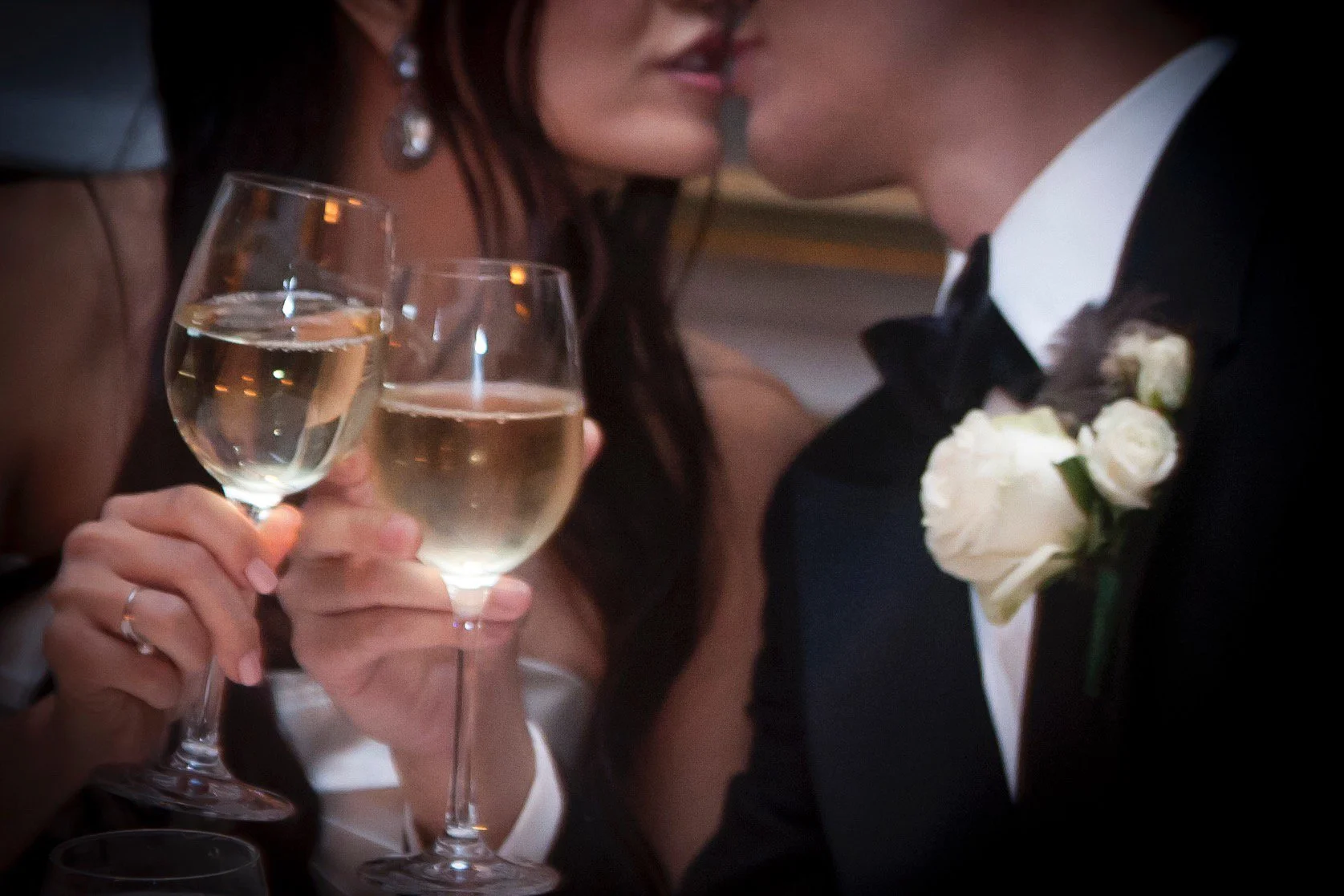 A close-up of a couple at a wedding toast, with two glasses of champagne; the woman is in a strapless dress wearing earrings, and the man is in a tuxedo with a white boutonniere.