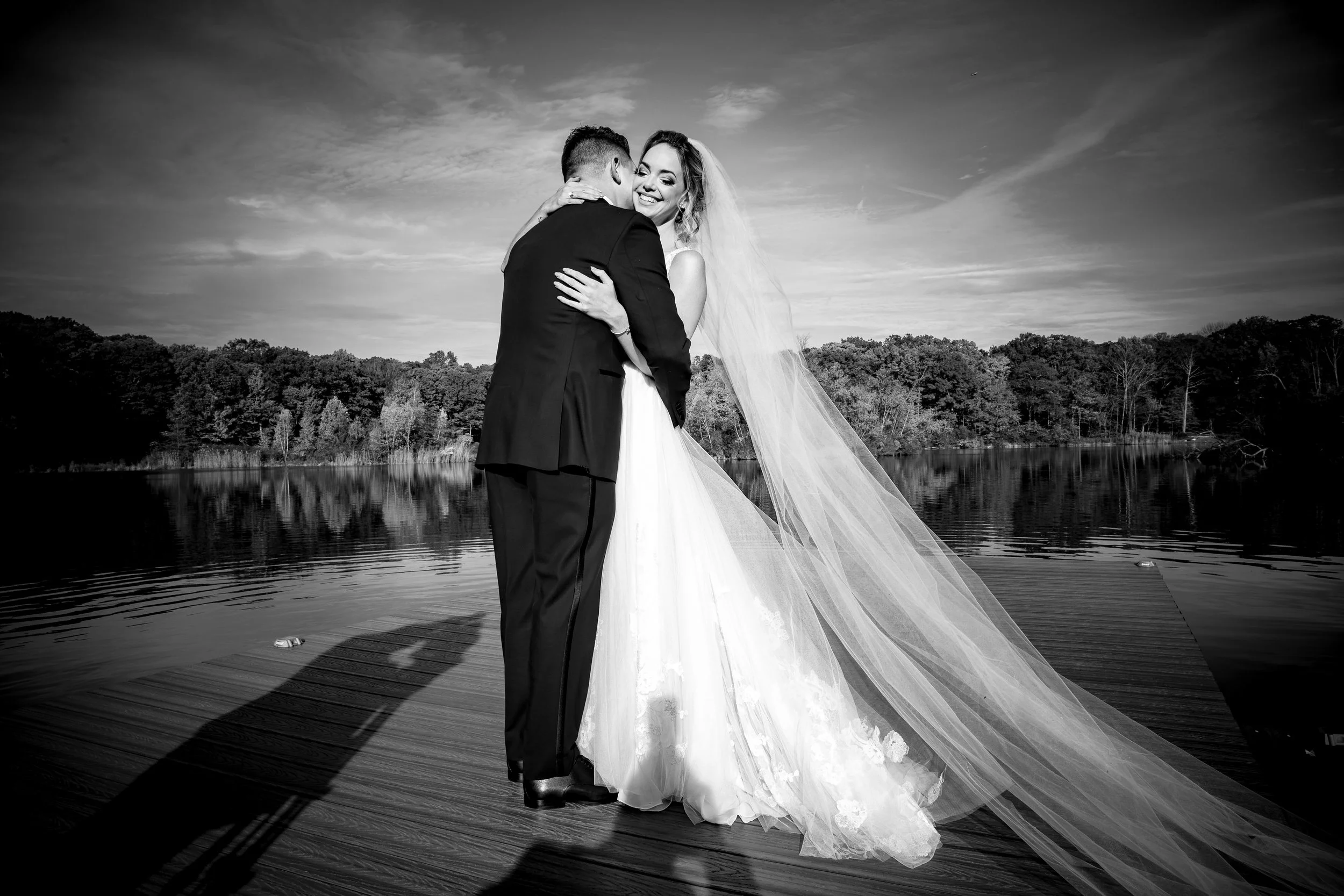 A bride and groom share a kiss on a dock by a lake during sunset, black and white photo.