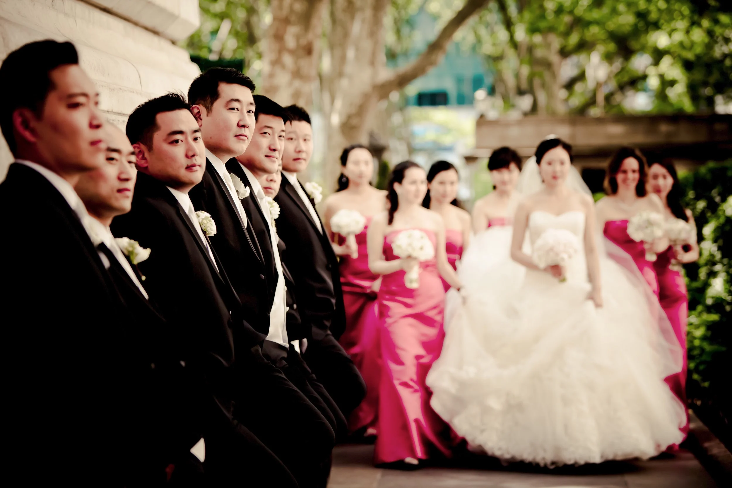A group of men in black tuxedos and women in pink and white dresses at a wedding ceremony outdoors, with trees in the background.