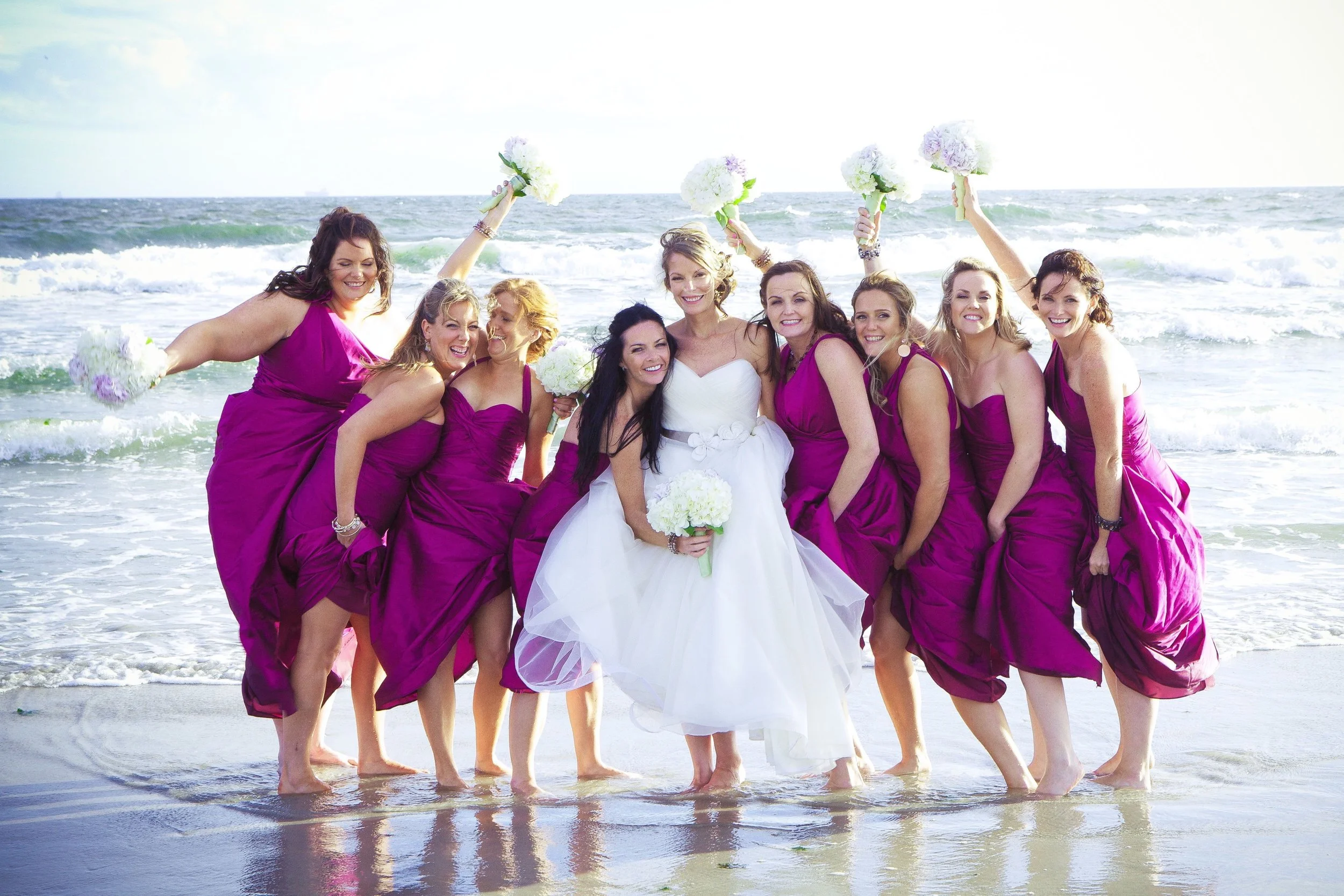A bride in a white wedding dress standing with her bridesmaids in purple dresses on the beach with waves in the background.