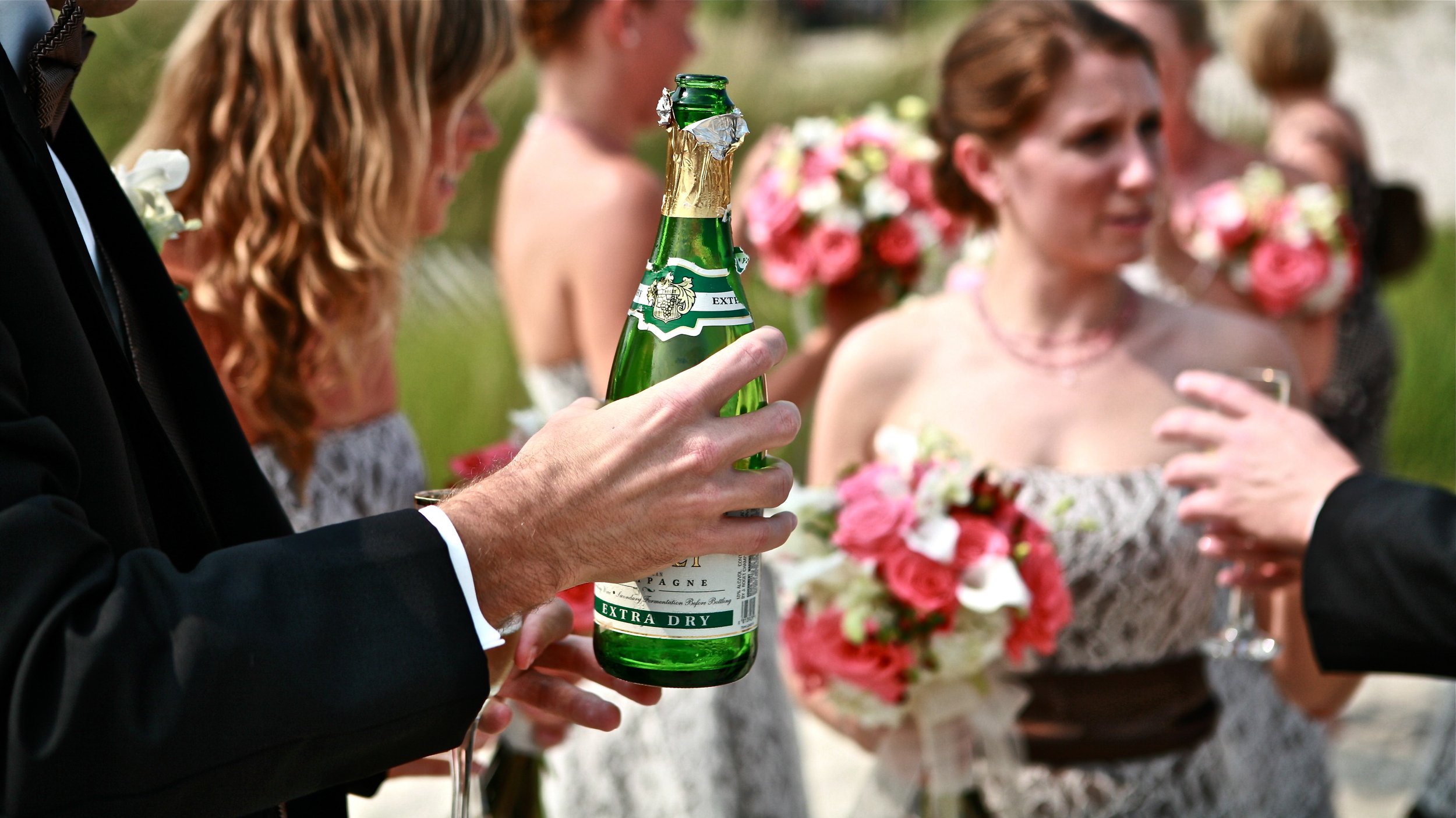 A person holding a green champagne bottle at a wedding reception with women in the background holding floral bouquets.