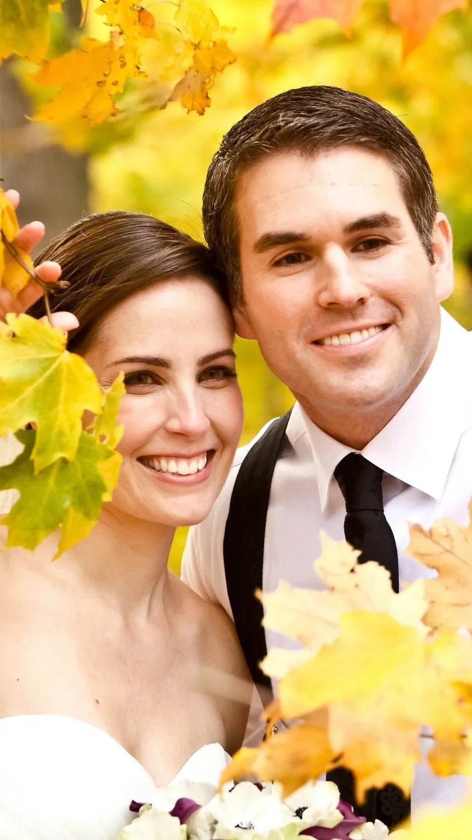 A smiling bride and groom taking a selfie outdoors during the fall, surrounded by colorful autumn leaves.