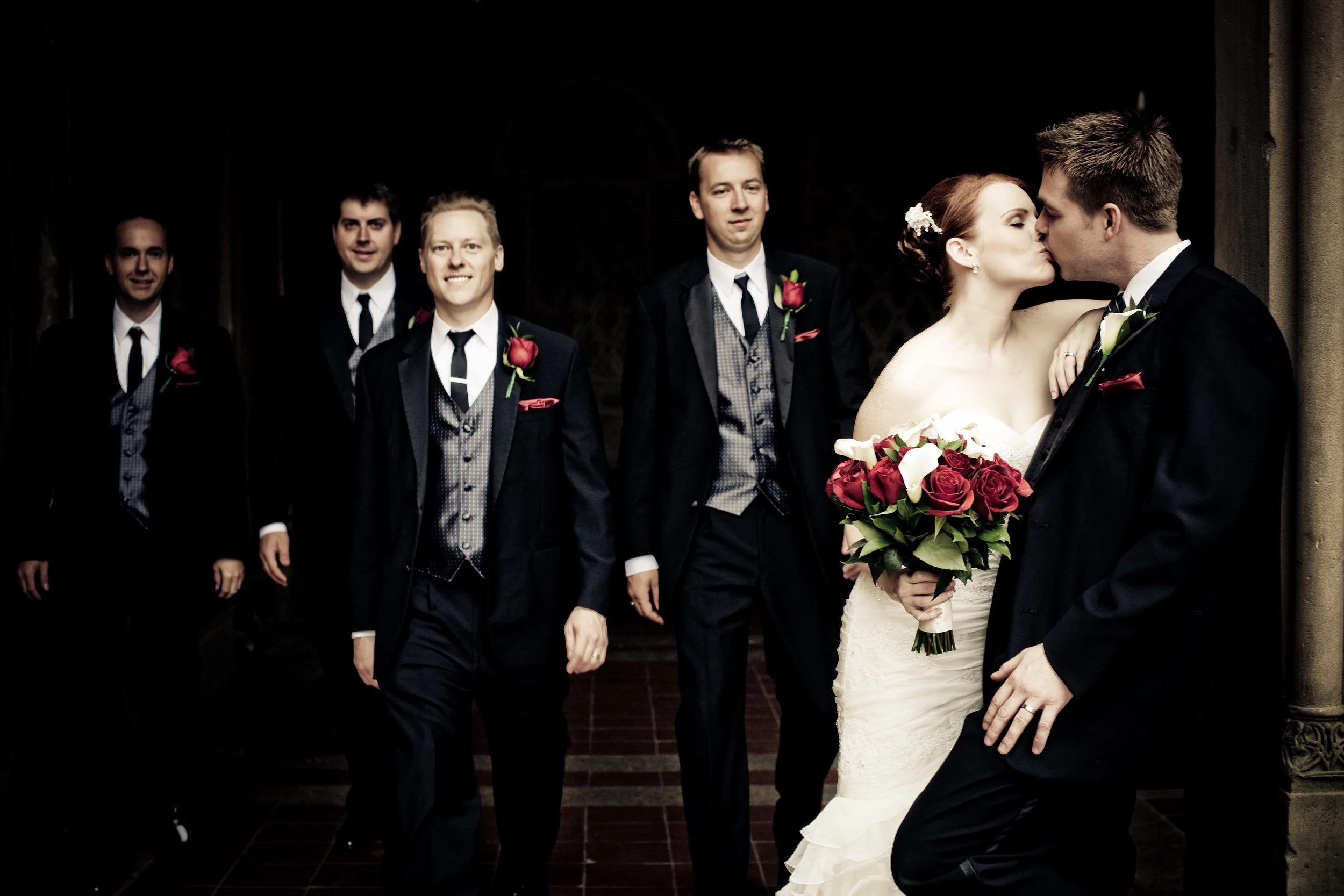 A bride and groom kissing, with the bride holding a bouquet of red and white roses, surrounded by four groomsmen in formal suits.