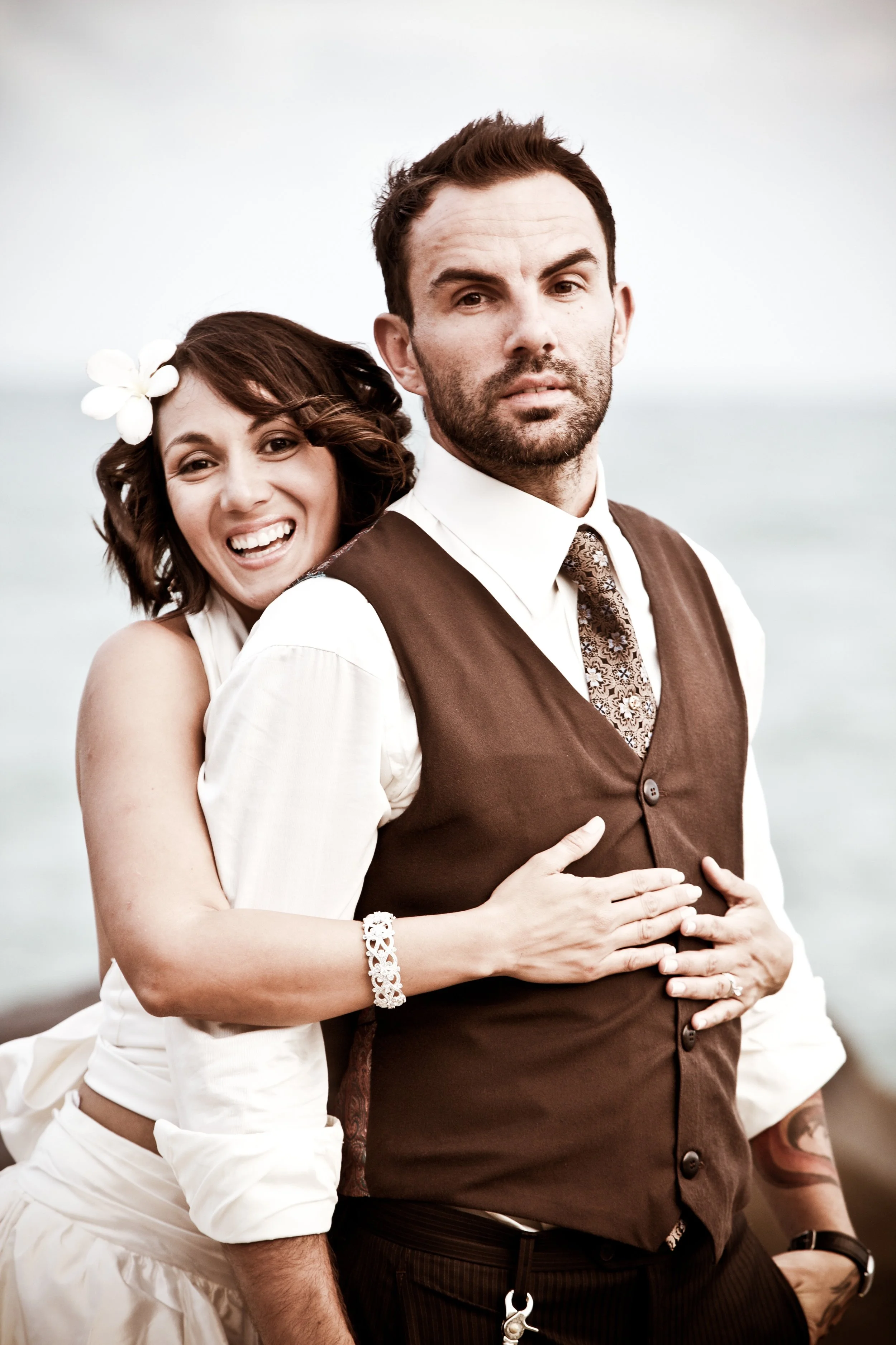 A smiling woman with brown hair and a white flower in her hair, hugging a serious-looking man with short dark hair and a beard, outdoors near the water.