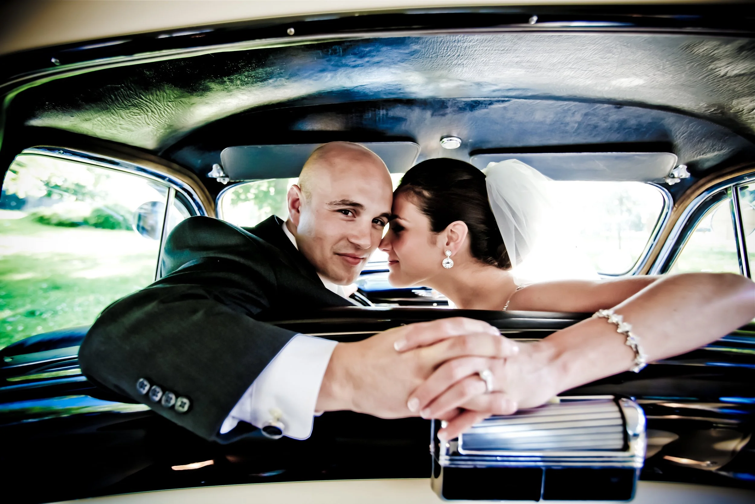 Newlywed couple in wedding attire sitting in a vintage car, leaning towards each other, smiling, with lush greenery visible through the windows.