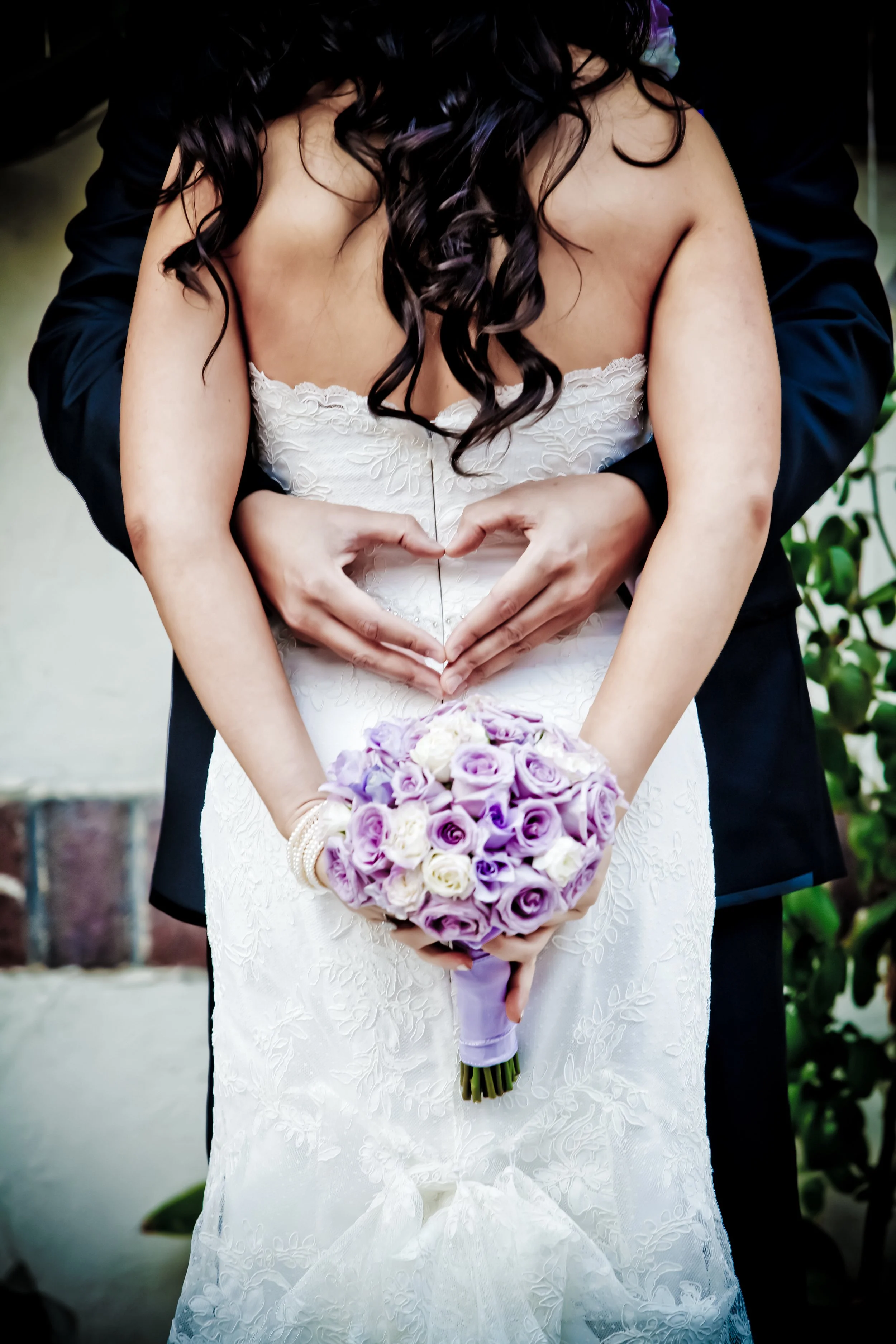A bride in a white wedding dress holding a bouquet of purple and white roses, with a person behind her forming a heart shape with their hands.