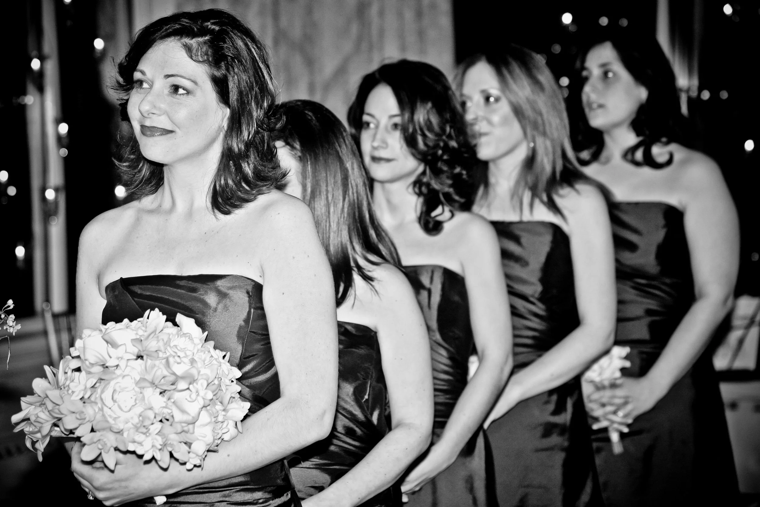 Black and white photo of five women standing in a line, dressed in strapless dresses at a formal event. The woman in the front holds a bouquet of flowers and is looking to her left. The women behind her have various hairstyles and are gazing forward 