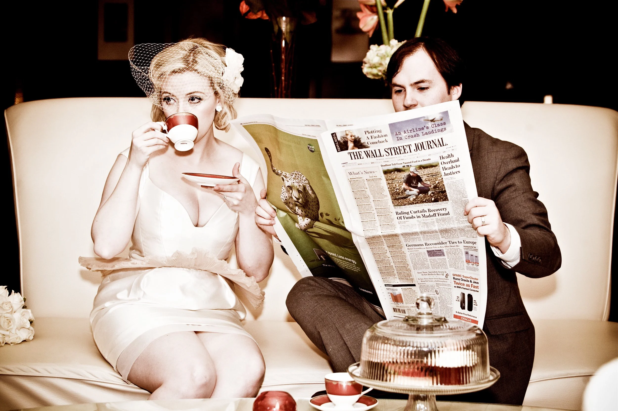 A woman in a white dress with a veil and flower headpiece drinks tea from a cup, while a man in a dark suit reads a newspaper titled 'The Wall Street Journal' on a sofa. A cake stand with a cake, a red and white teacup and saucer, and a small red orn