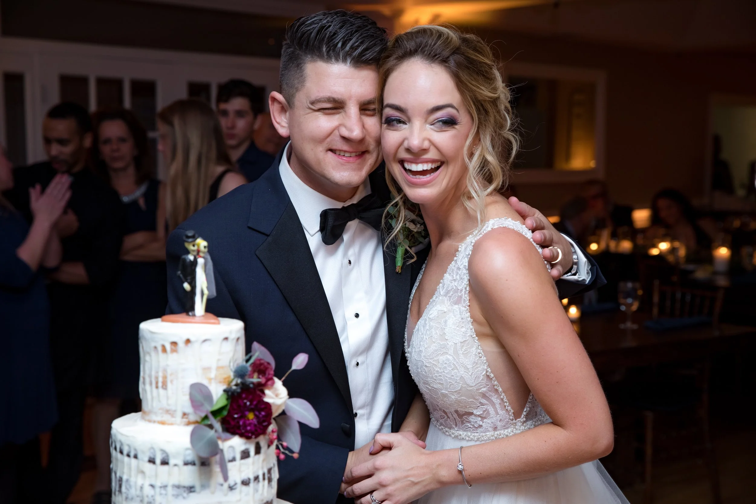 Bride and groom smiling and hugging at their wedding reception, with a wedding cake decorated with flowers and a bride and groom cake topper.