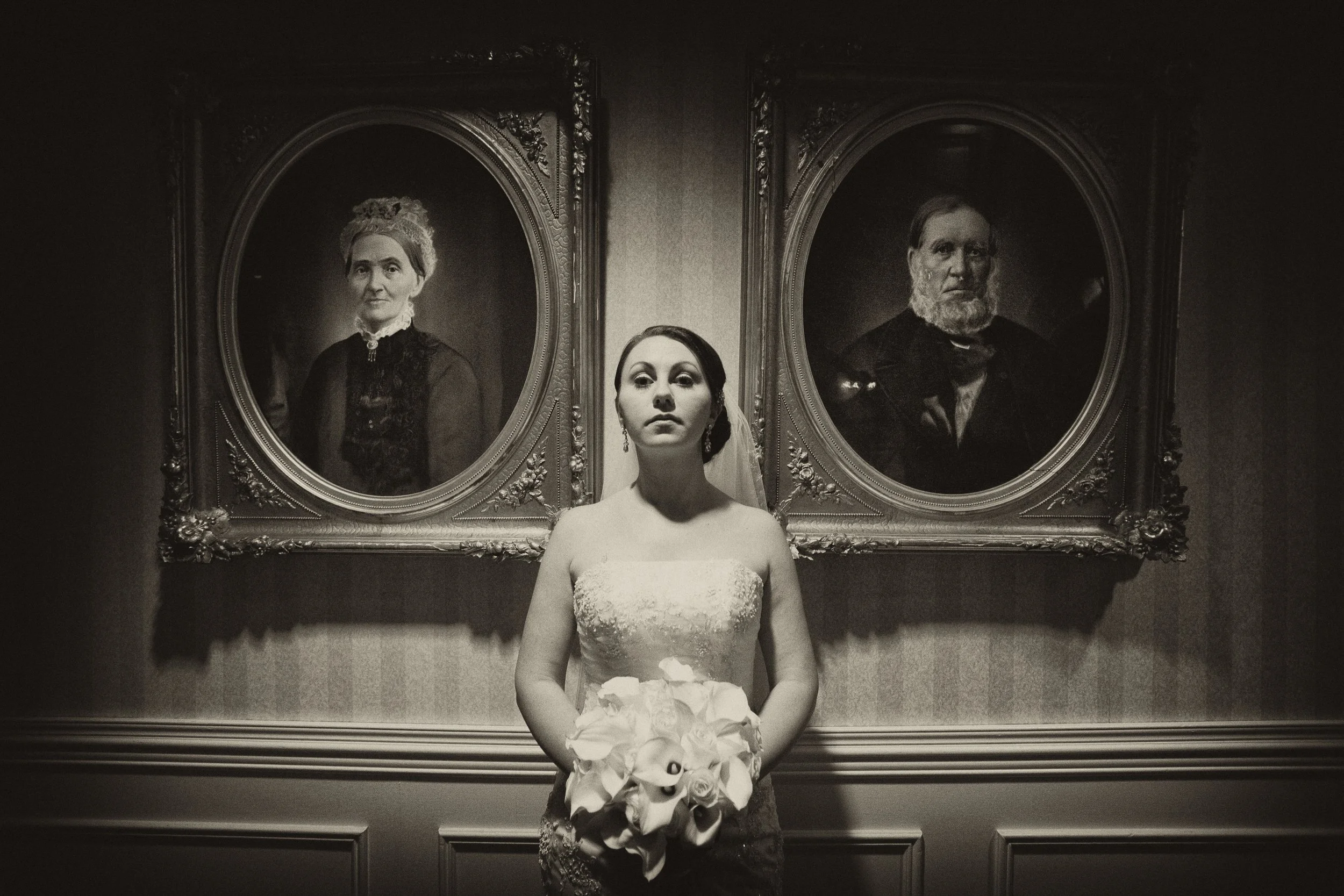 A woman in a wedding dress holding a bouquet of calla lilies standing in front of two framed portraits on a wall.