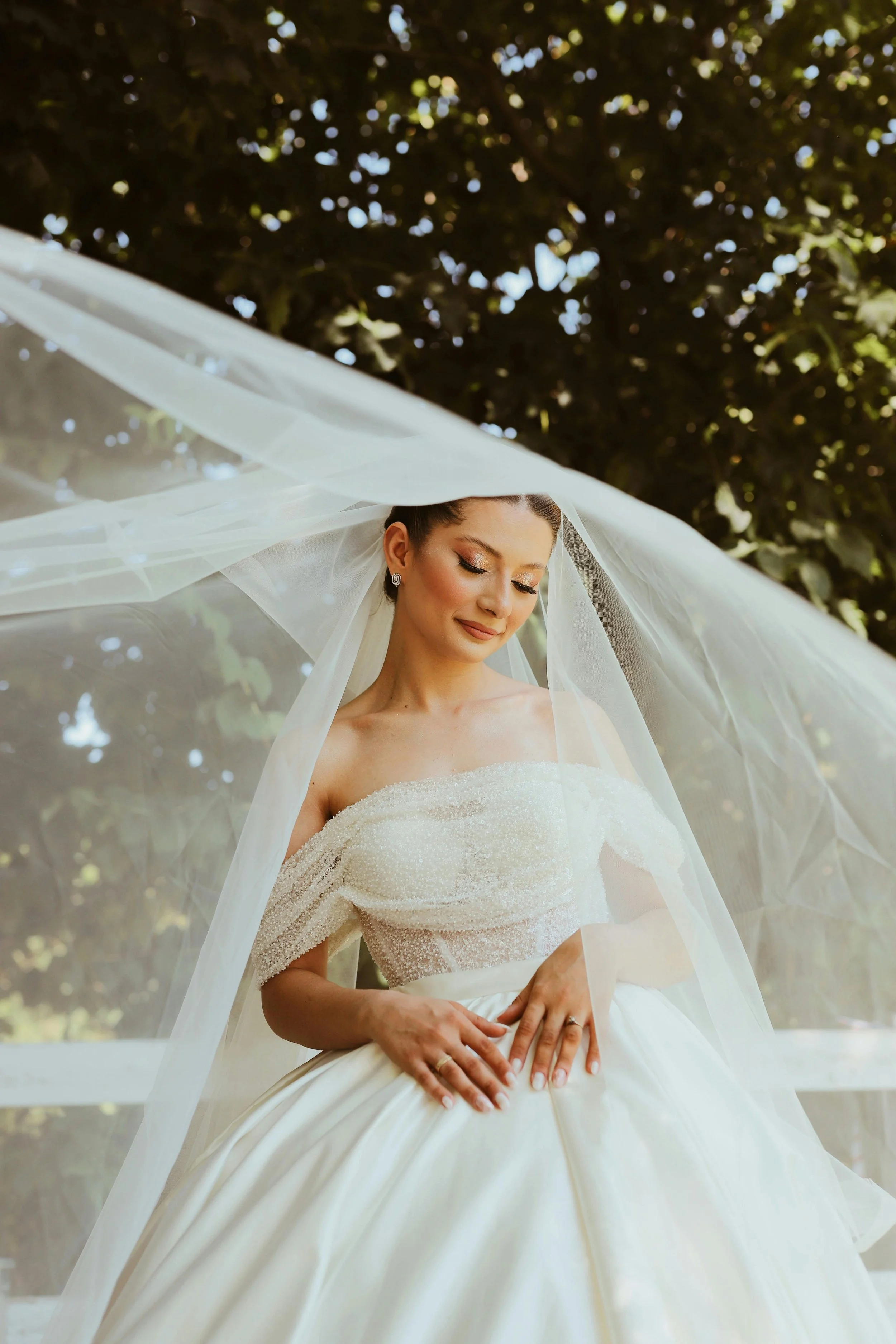 A bride in a white wedding gown and veil standing outdoors with a background of trees, looking down with a serene expression.