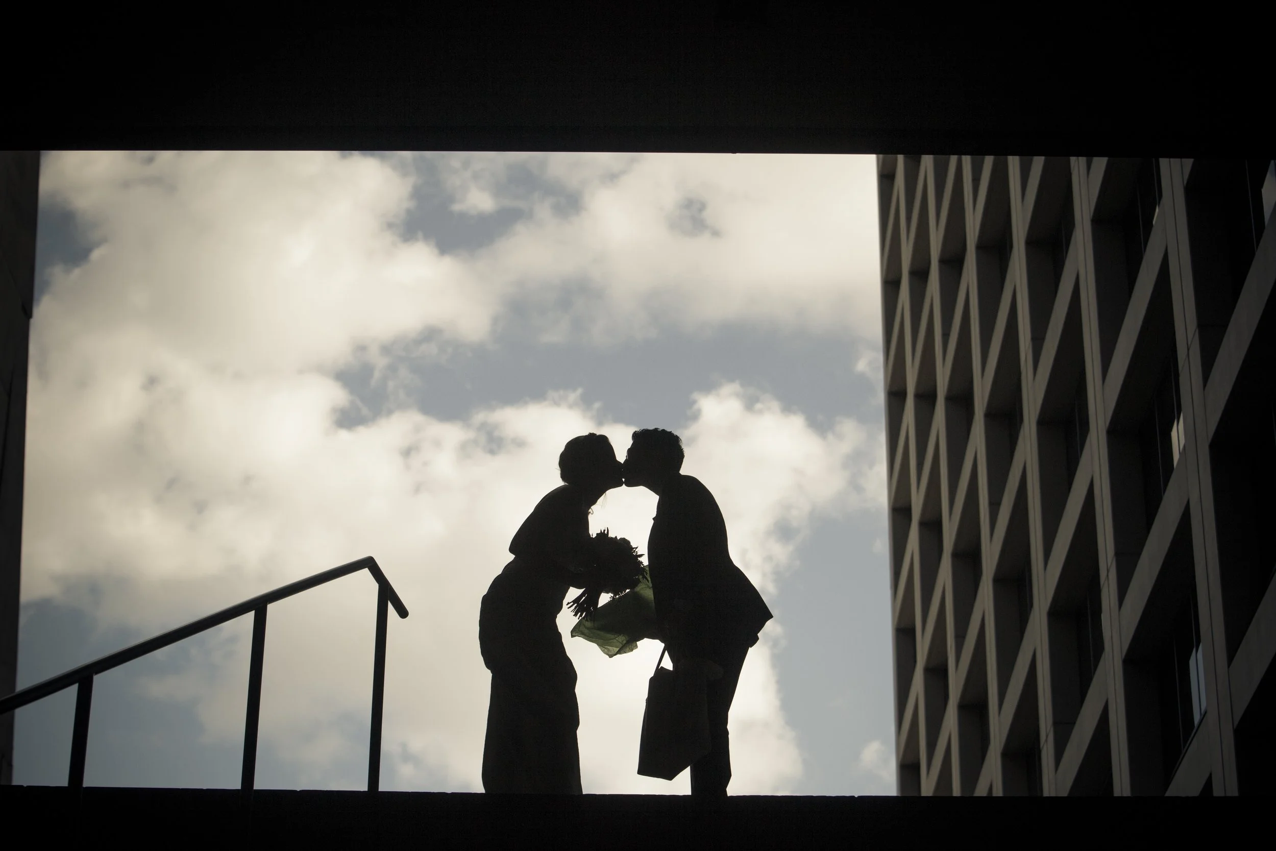 Silhouette of a couple kissing, one holding a bouquet of flowers, on a balcony overlooking the cloudy sky, with modern building on the right side.