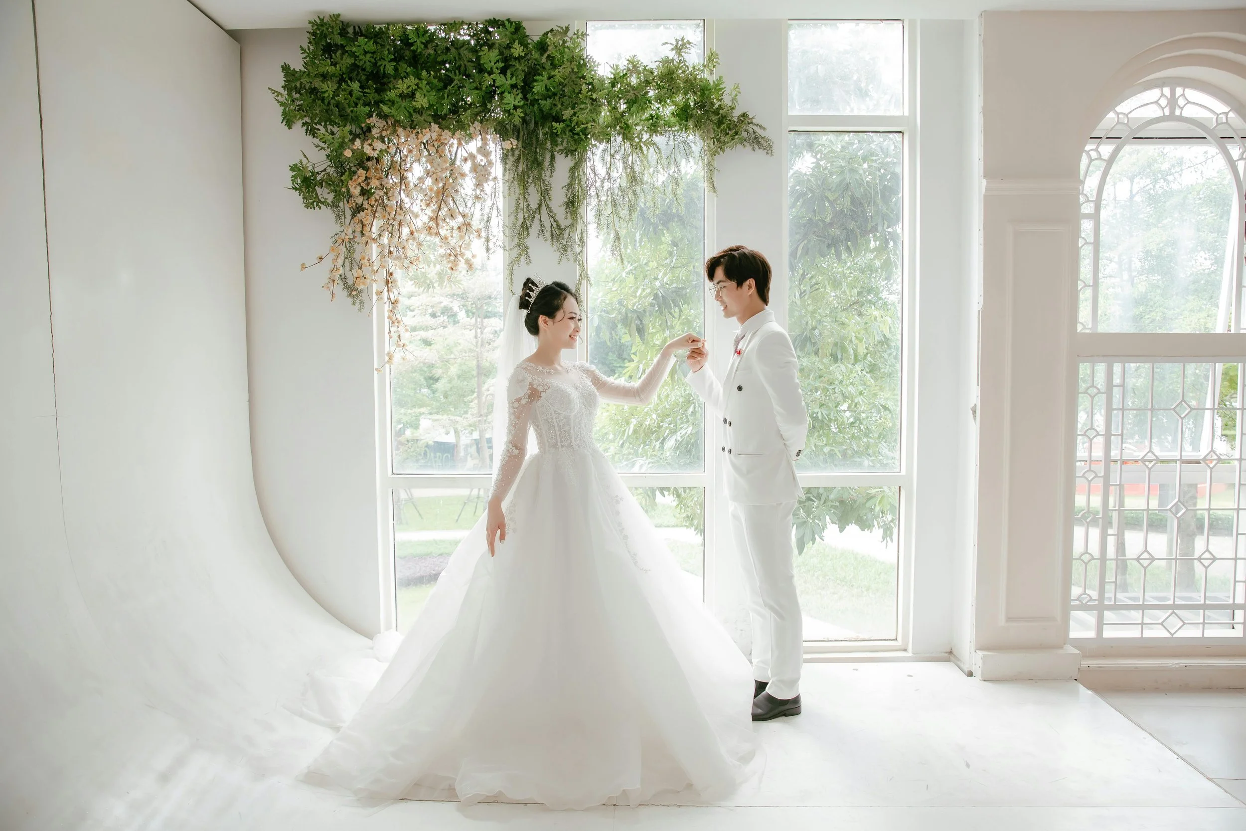 A bride and groom in wedding attire holding hands and smiling indoors near large windows with trees outside.