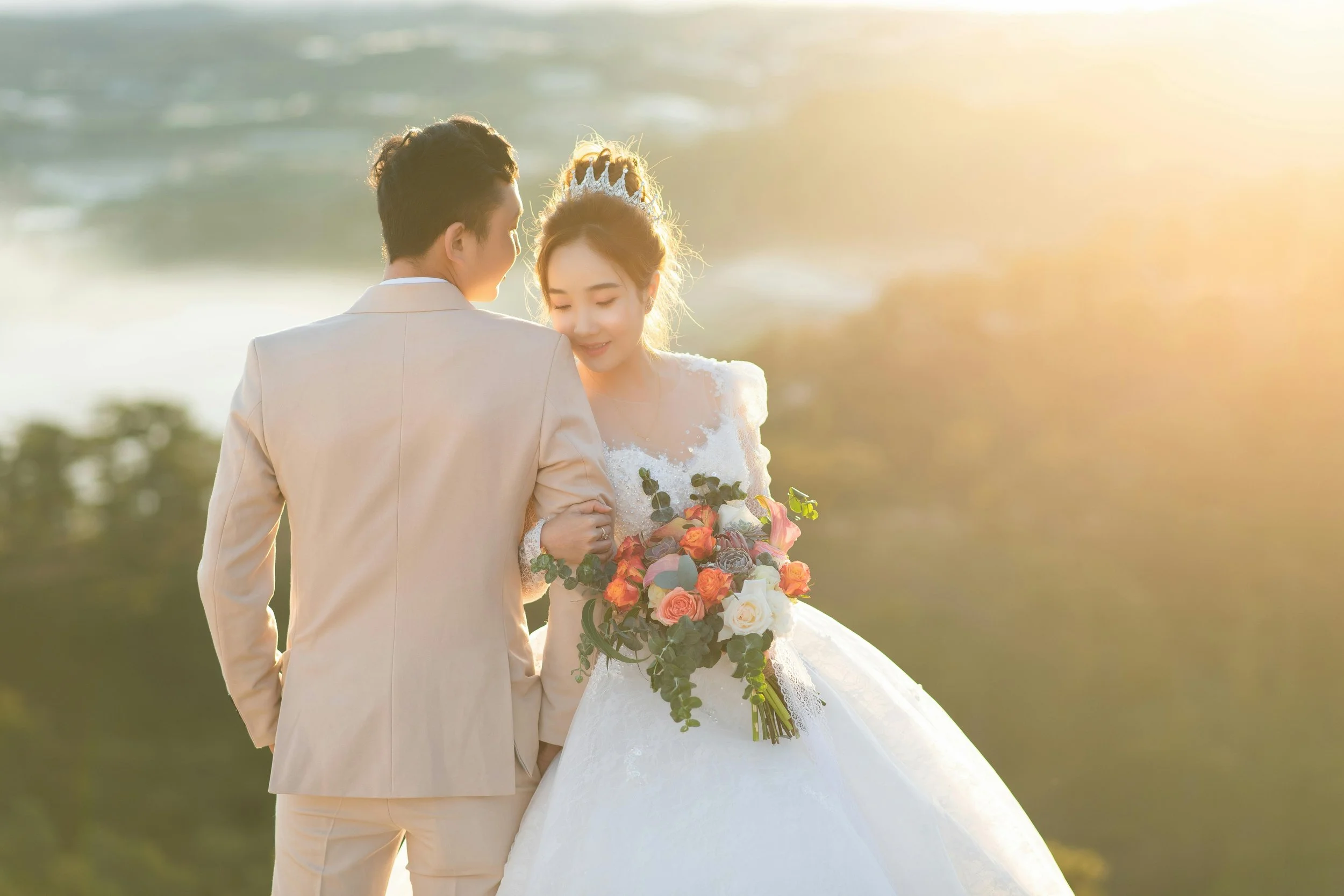 A bride in a wedding dress holding a bouquet of flowers and an emotional groom dressed in a beige suit, with a scenic outdoor background and warm sunlight.