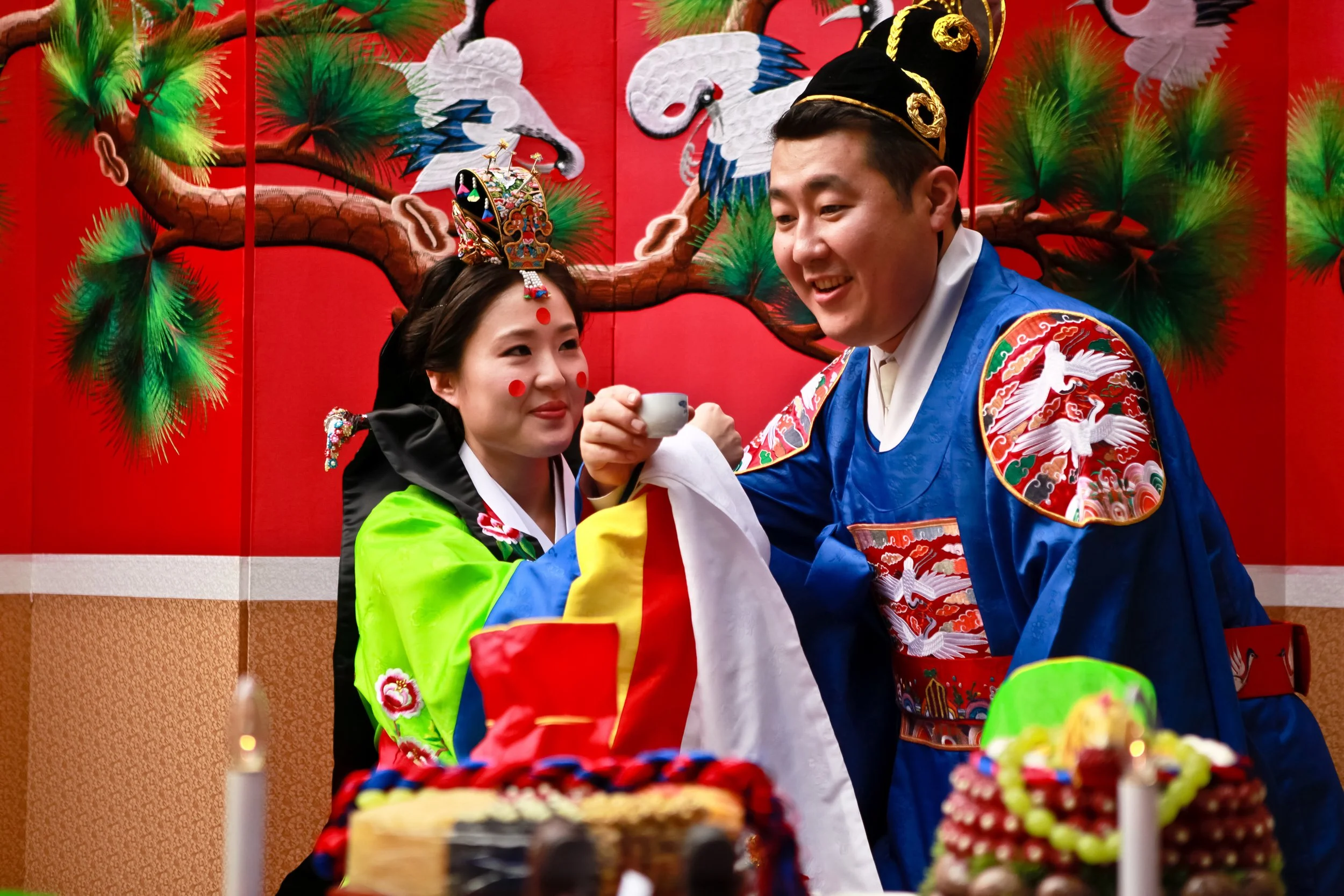 A couple dressed in traditional Asian attire during a celebration. The woman is wearing a brightly colored dress with a crown and face decorations, and the man is in a blue robe with embroidery and a headdress. They are smiling, holding a small tea c