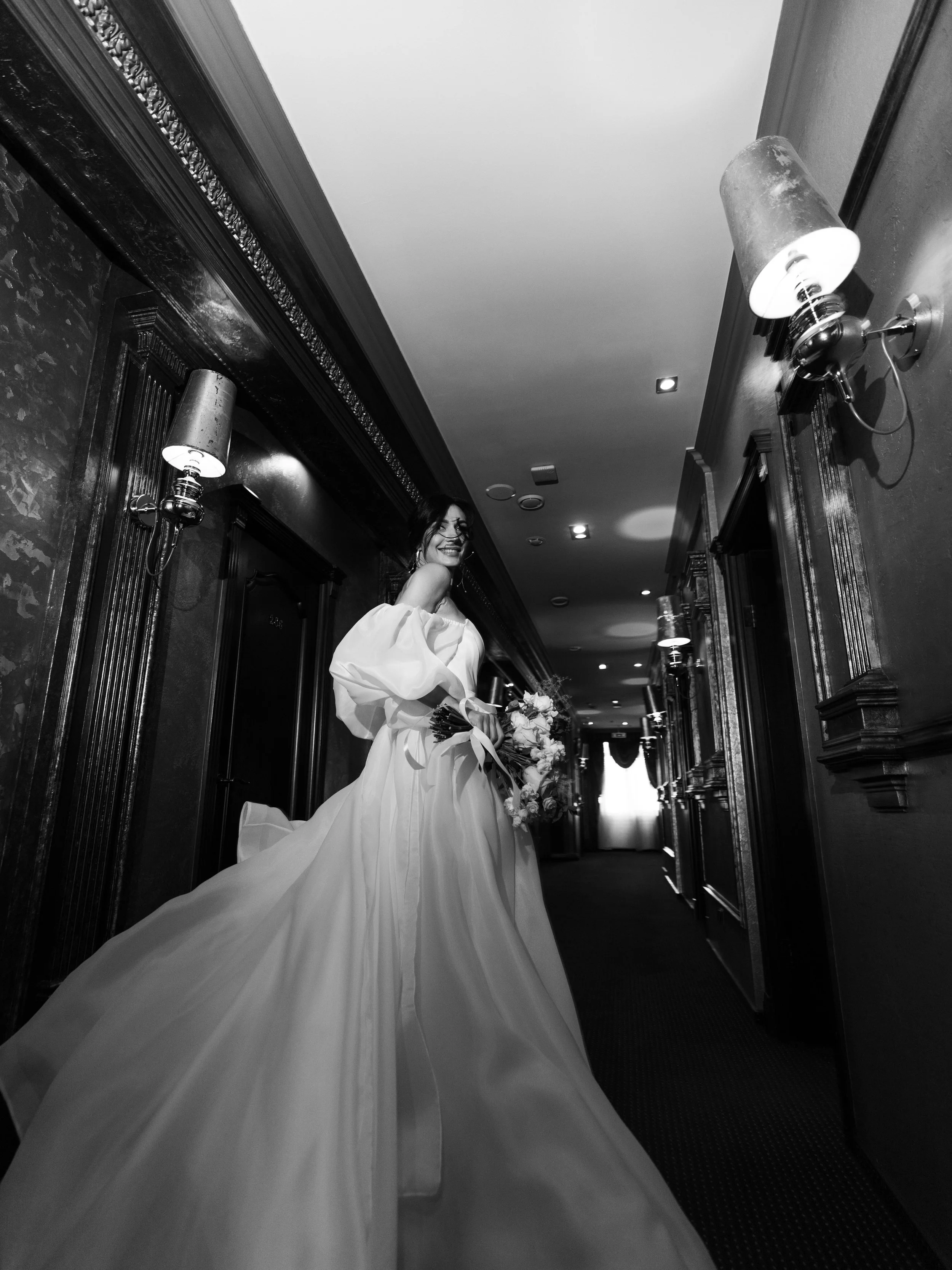 A woman in a wedding dress holding a bouquet, standing in a dimly lit hallway with ornate wood paneling and wall sconces, smiling.