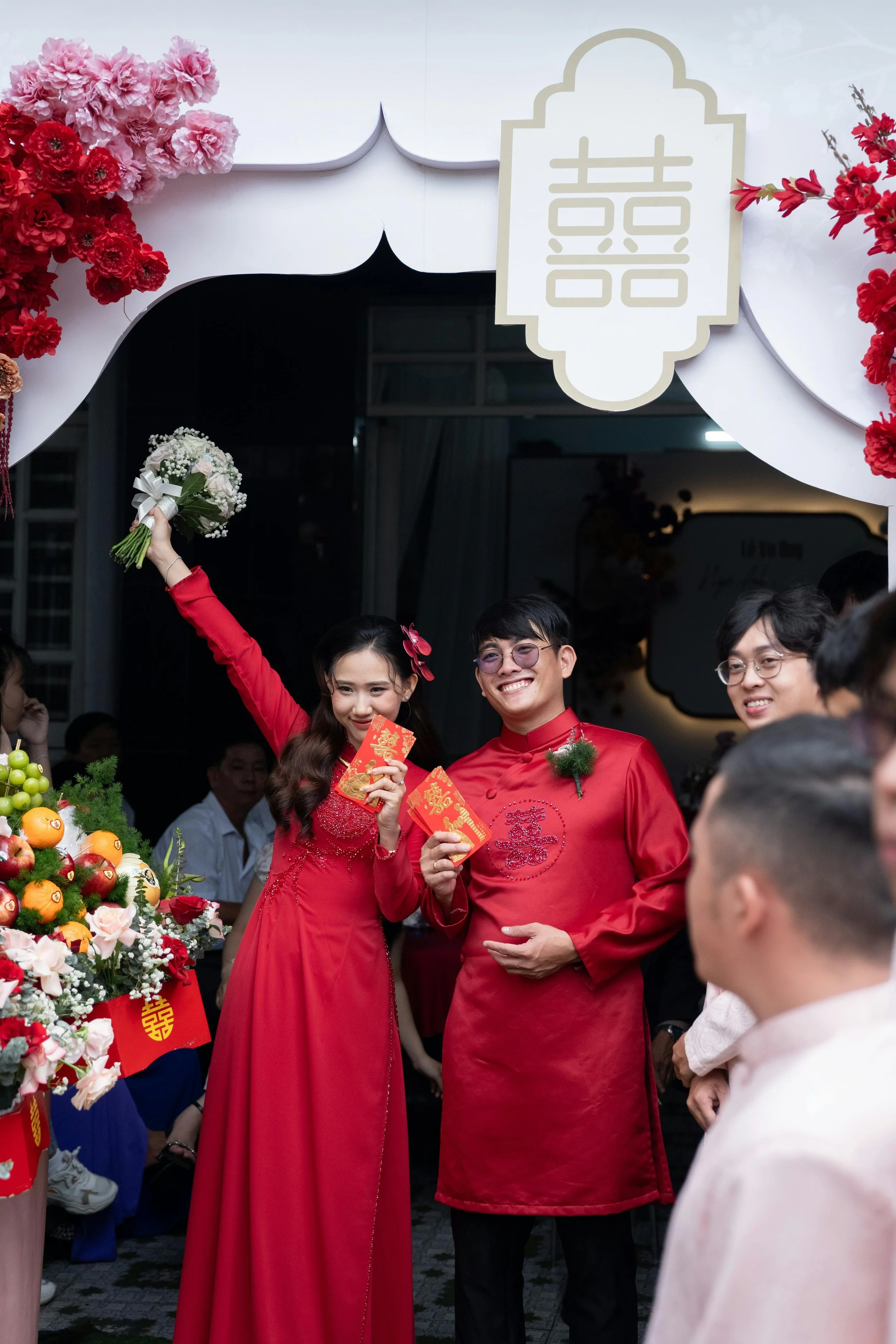 Couple celebrating their wedding, dressed in red traditional Vietnamese attire, holding red envelopes and a bouquet, surrounded by friends, flowers, and wedding decorations.