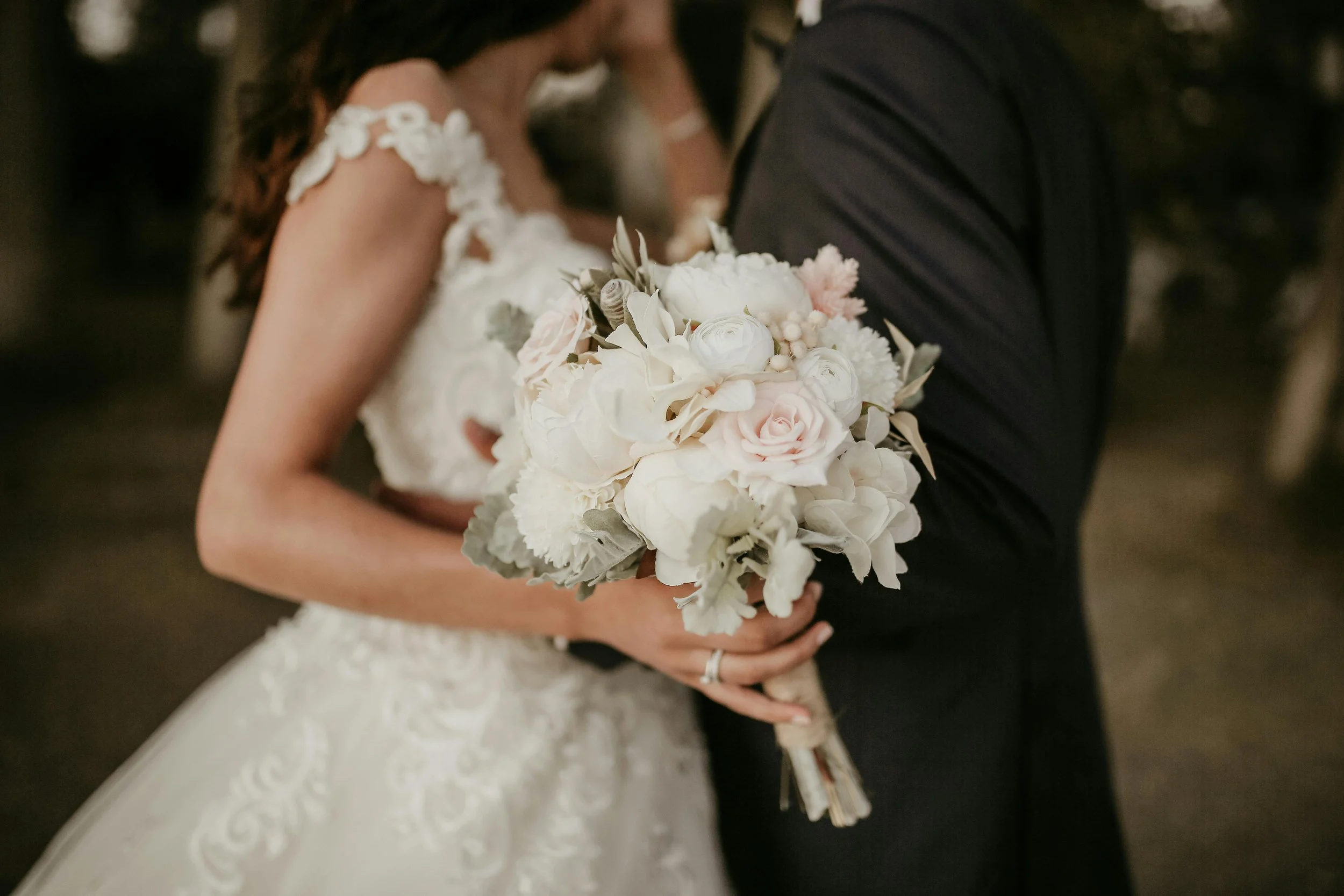 A bride and groom holding a bouquet of white and blush pink flowers, with the bride wearing a lace wedding dress and the groom in a black suit.