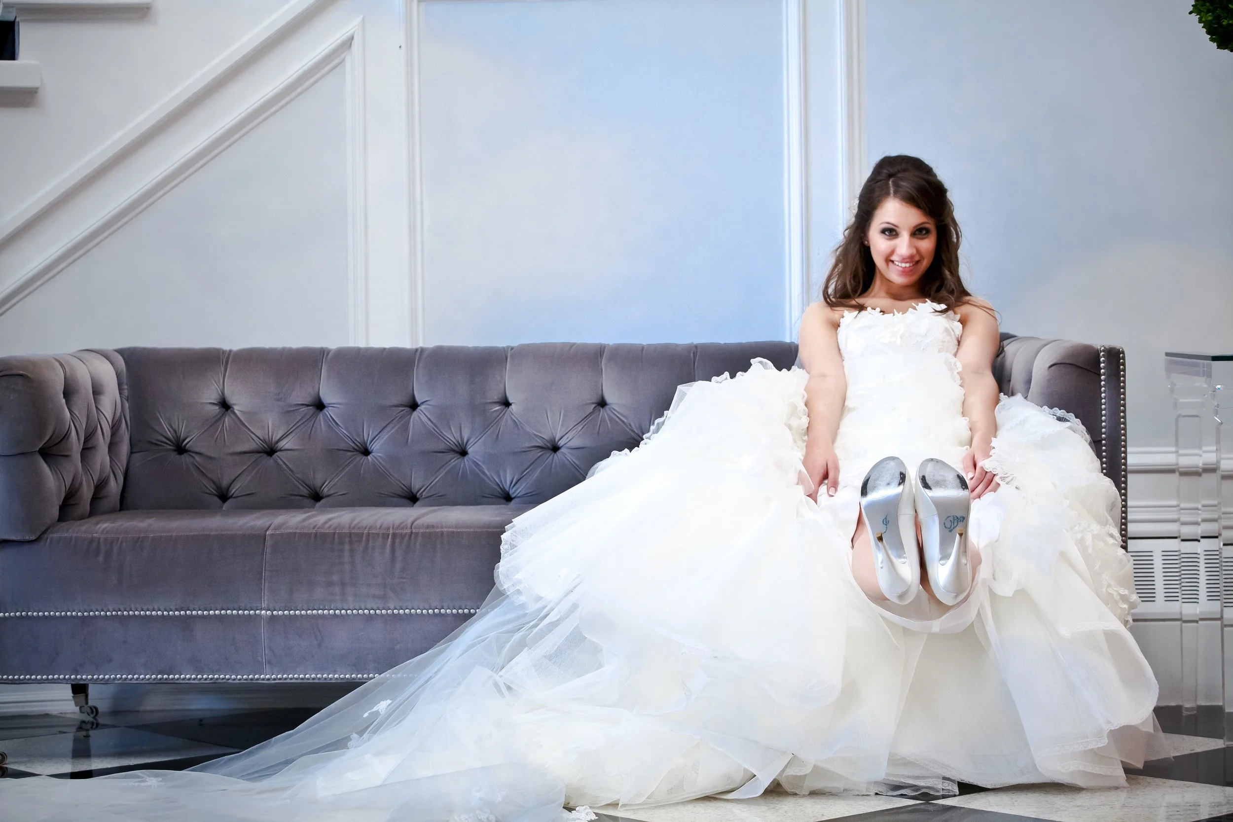 A young woman in a white wedding dress sitting on a gray velvet sofa, lifting her wedding gown to reveal white shoes, smiling in a bright indoor space.