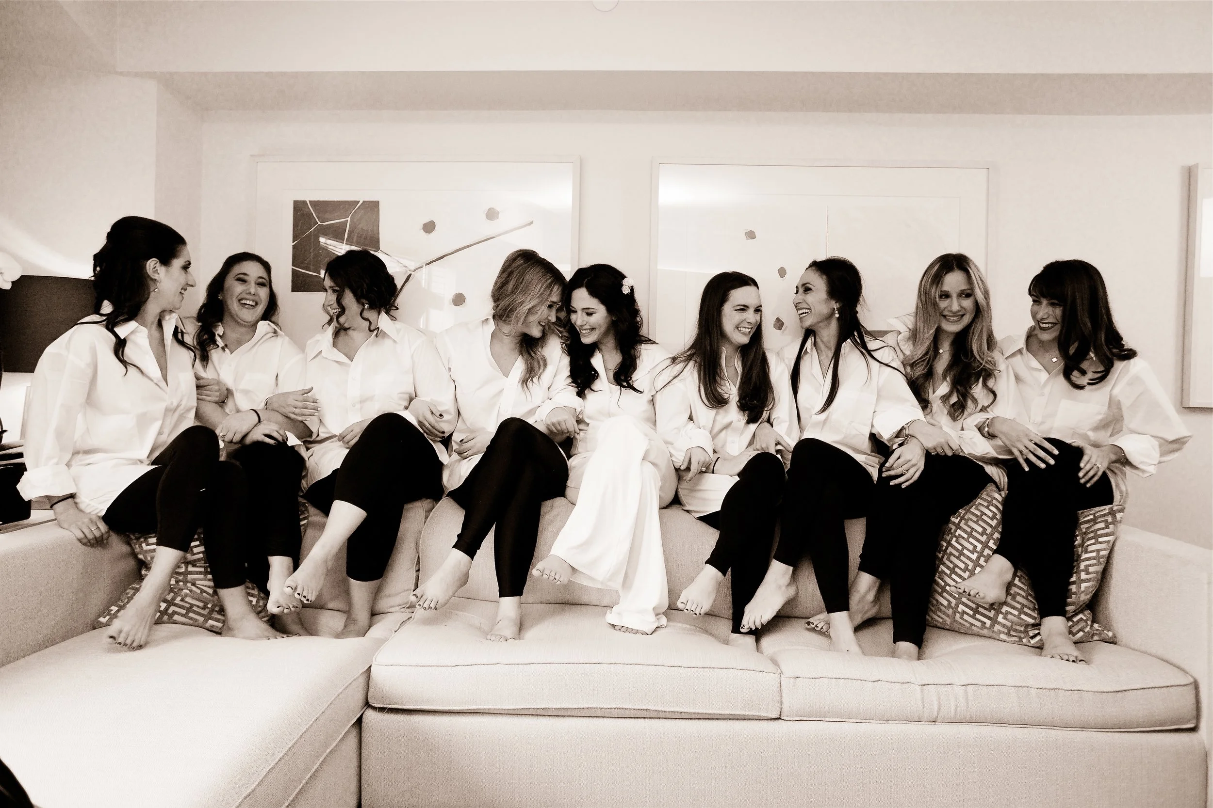 Group of women dressed in white shirts, sitting on a couch, laughing and enjoying each other's company.