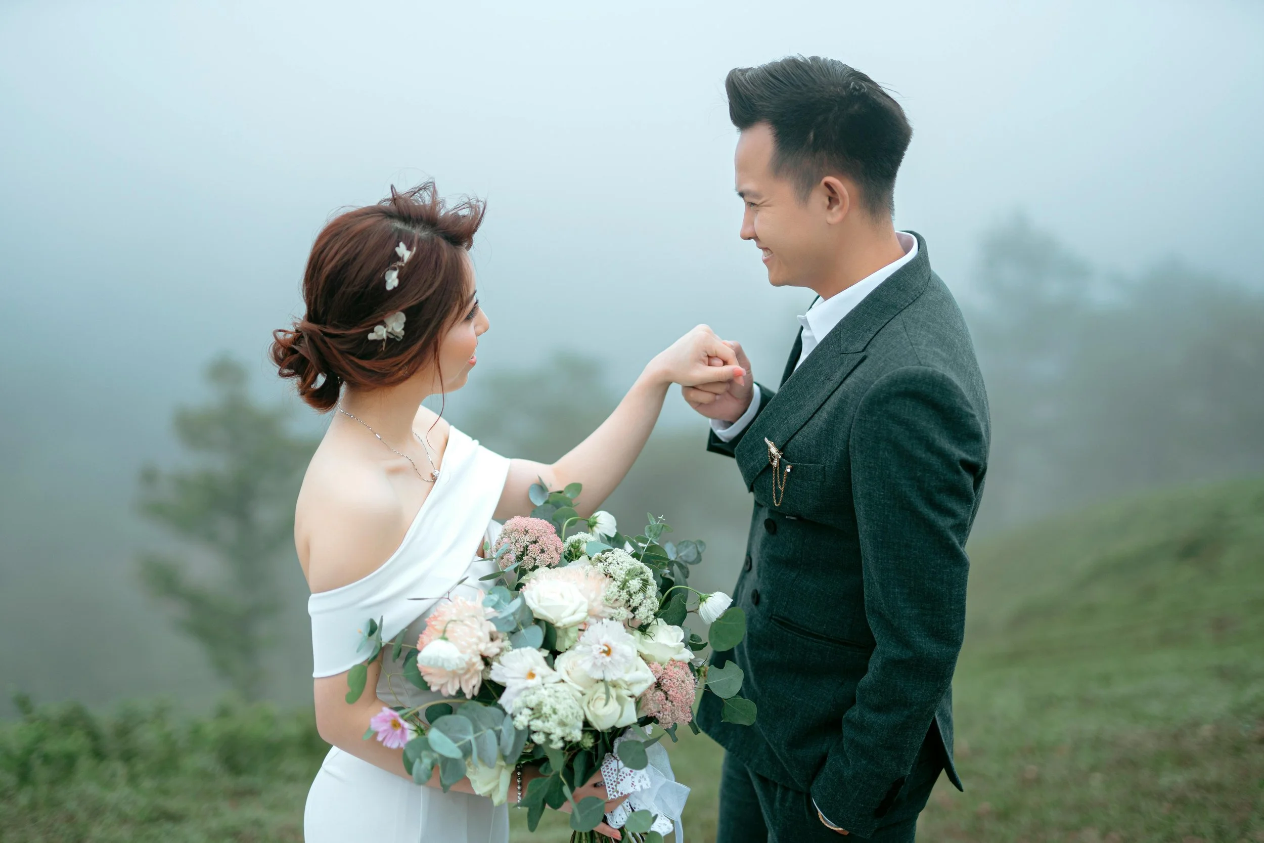 A bride and groom smiling and holding hands outdoors in a foggy, green landscape. The bride wears a white off-shoulder dress with floral accessories in her hair and holds a large bouquet of flowers. The groom is dressed in a gray suit with a white sh