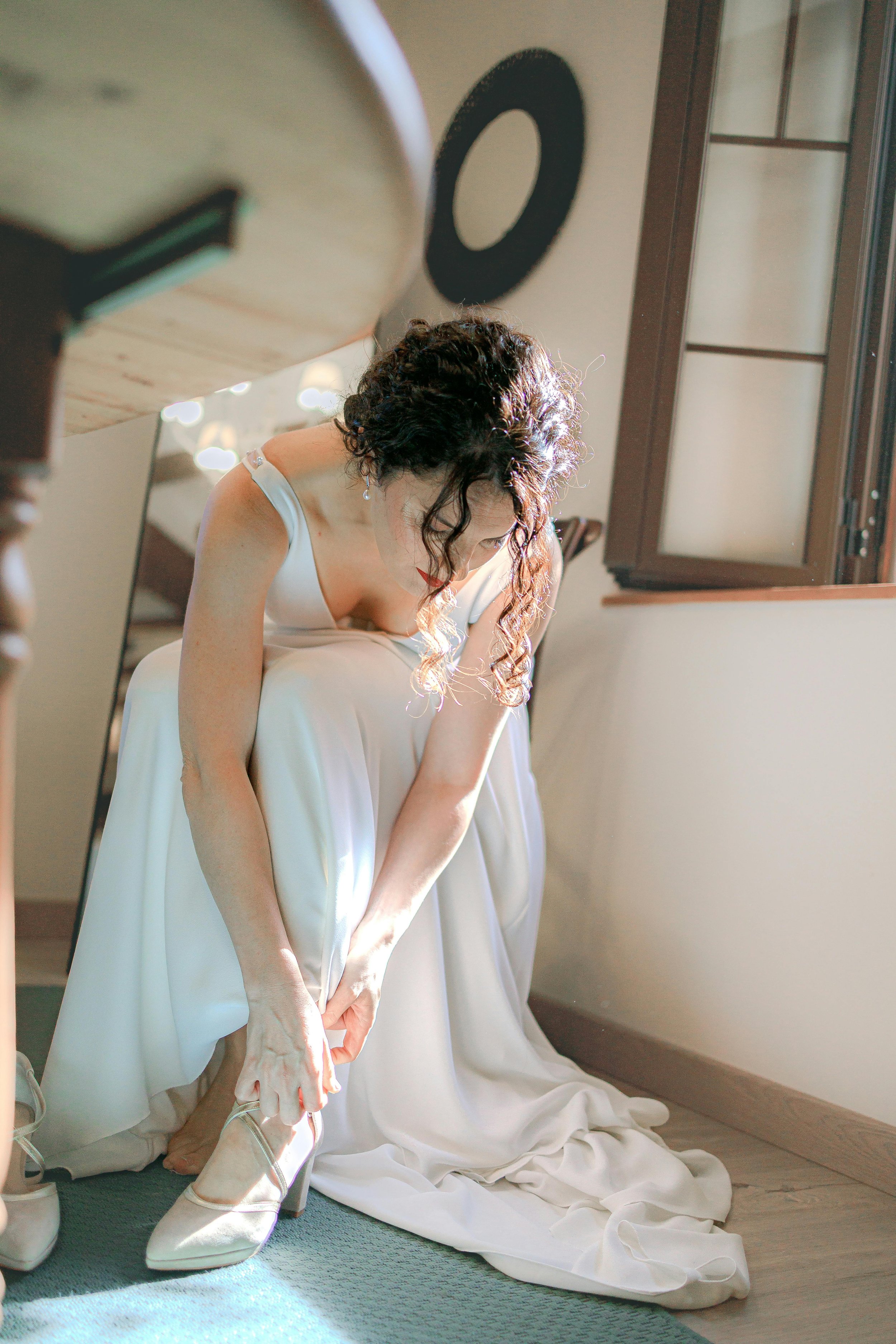 A woman in a white dress kneels on the floor, adjusting her high heel shoe as she prepares for her wedding. She is in a room with a wooden floor and open window, with sunlight casting light on her and the surroundings.