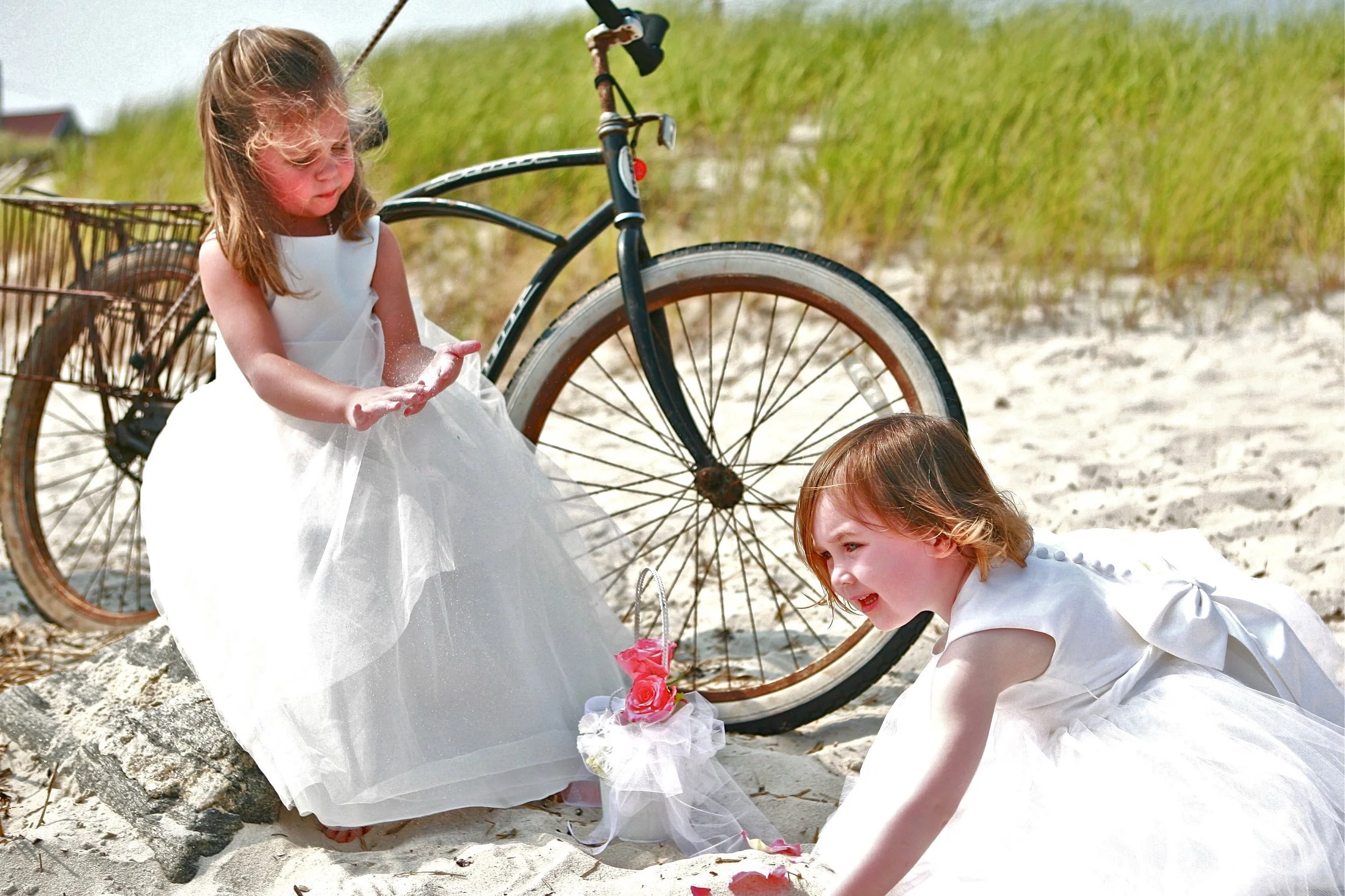 Two young girls in white dresses playing in the sand near a bicycle with a basket, on a beach with grassy dunes in the background.