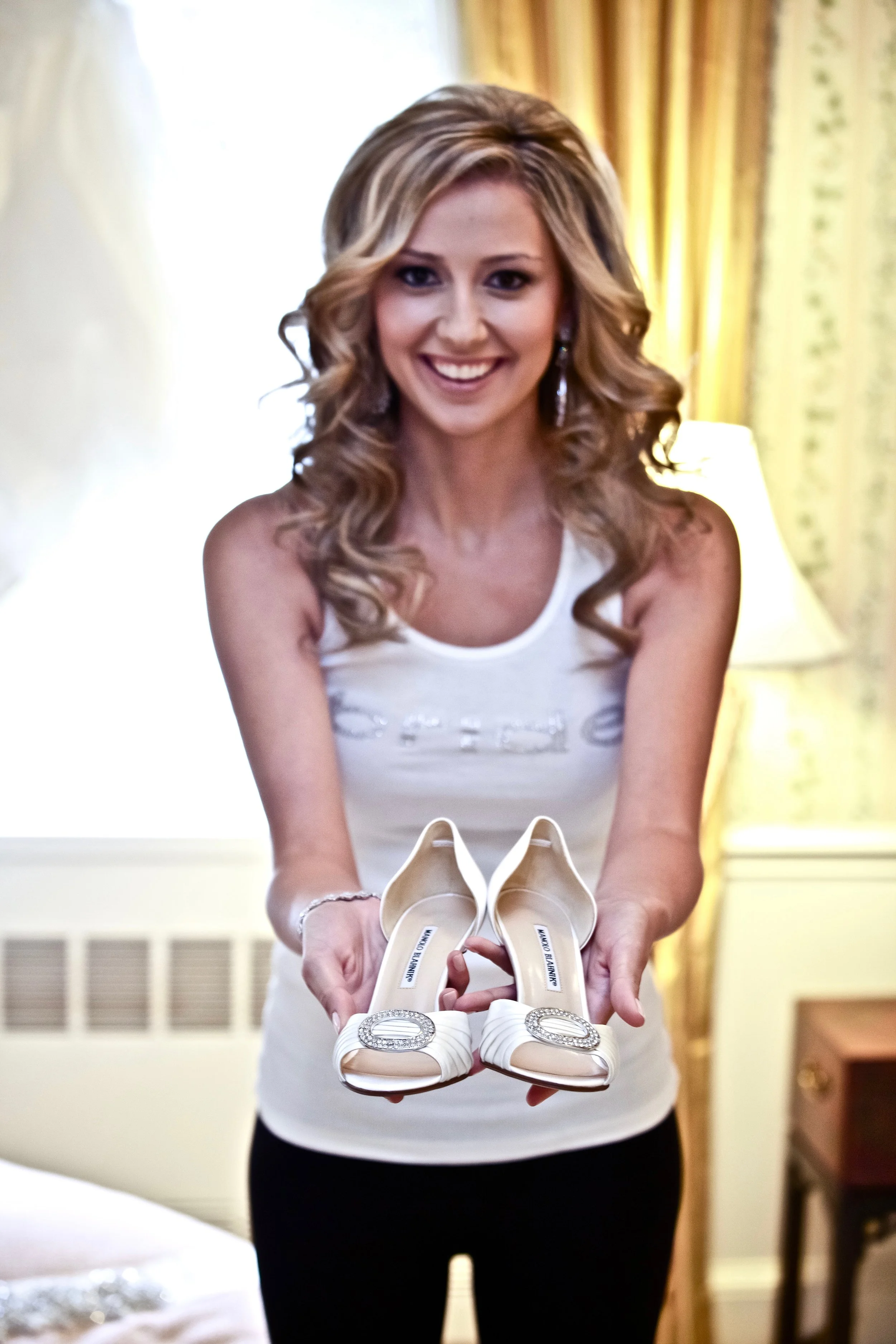 A woman with blonde, curly hair smiling and holding a pair of white high-heeled shoes with decorative buckles.