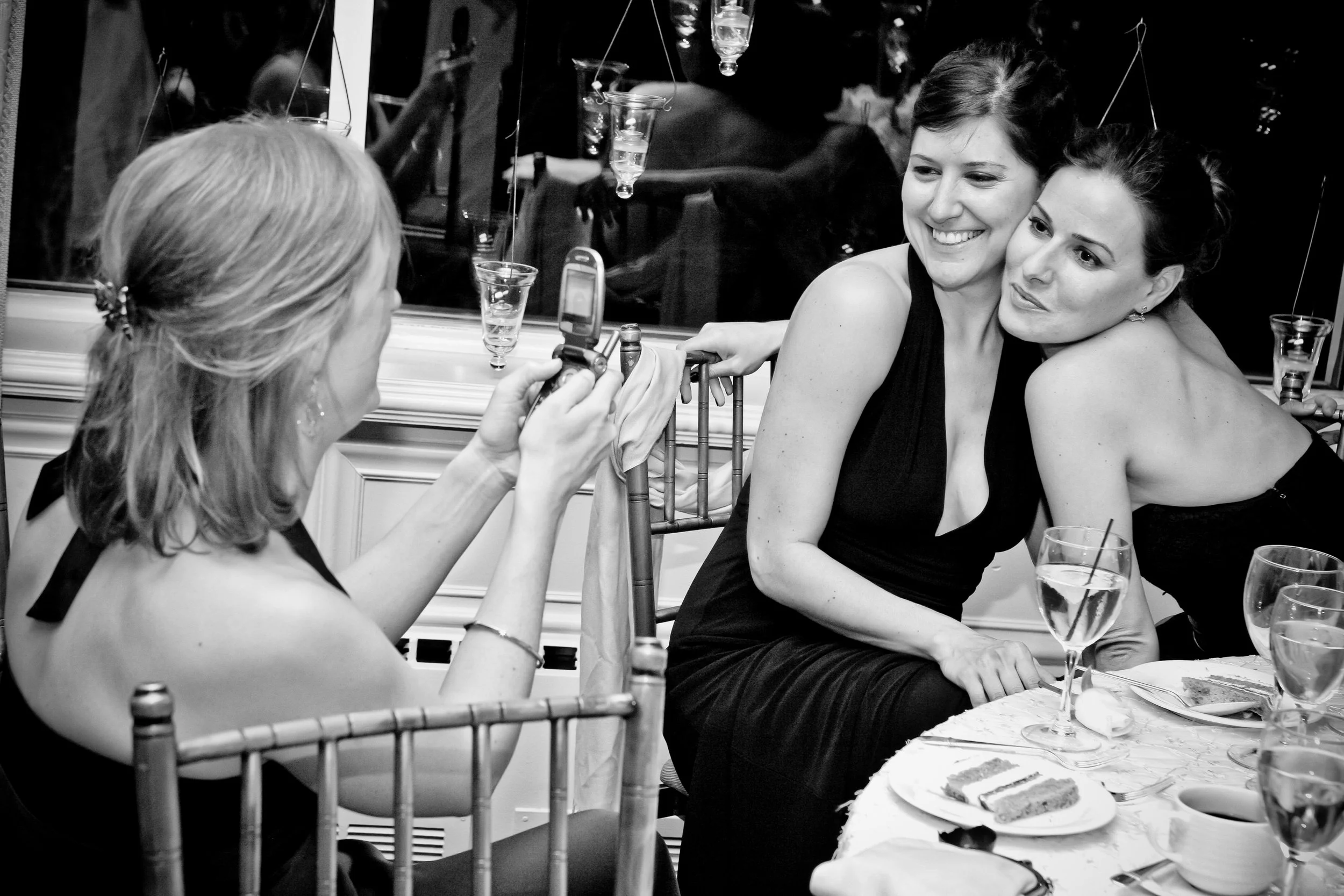 Two women dressed in black at a dinner table taking a photo of another woman, who is also dressed in black, with a flip phone. The table has glasses, plates with cake, and a tablecloth. The women are smiling and leaning close together.
