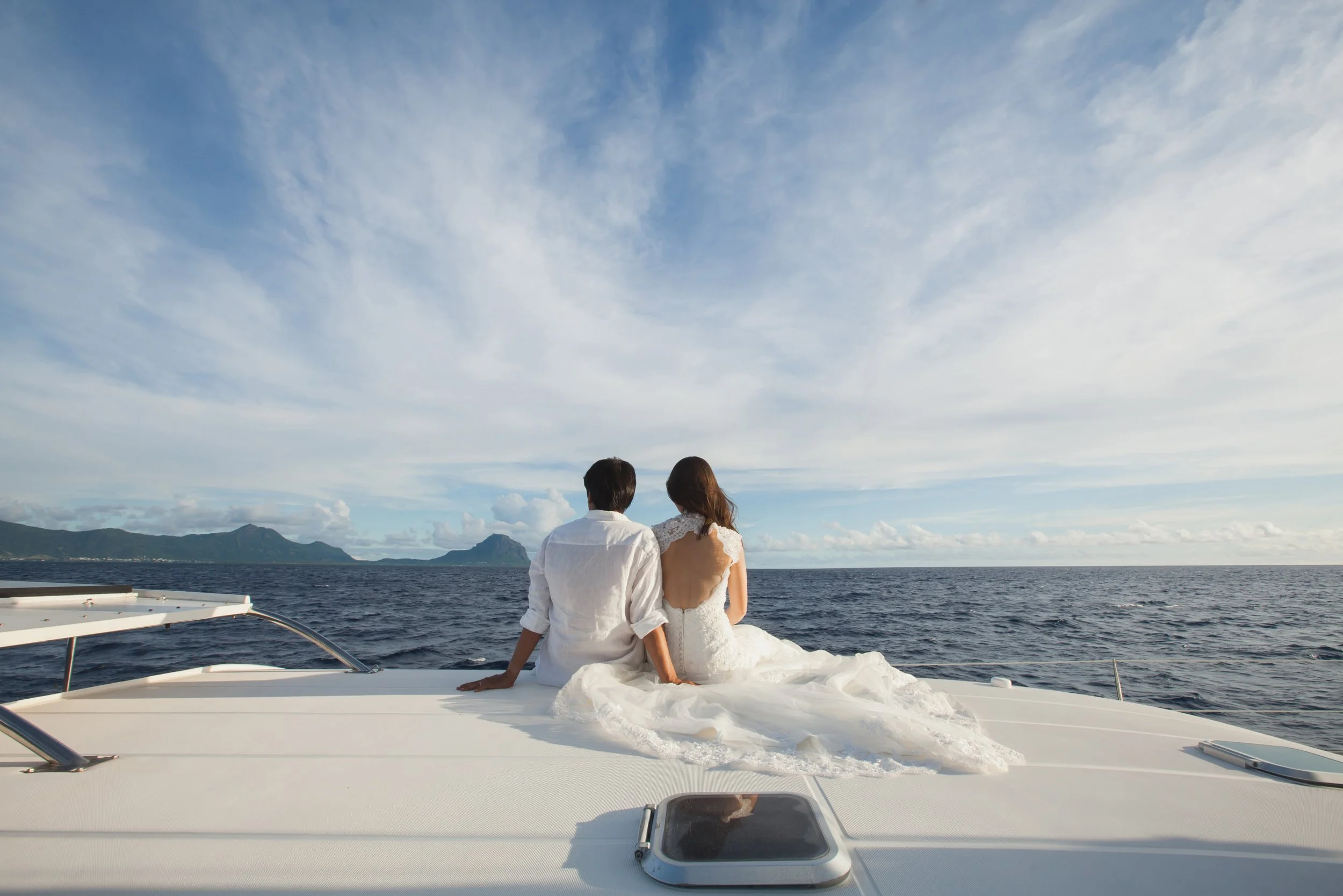 A couple in wedding attire sitting on the deck of a boat, facing the ocean with mountains in the distance.