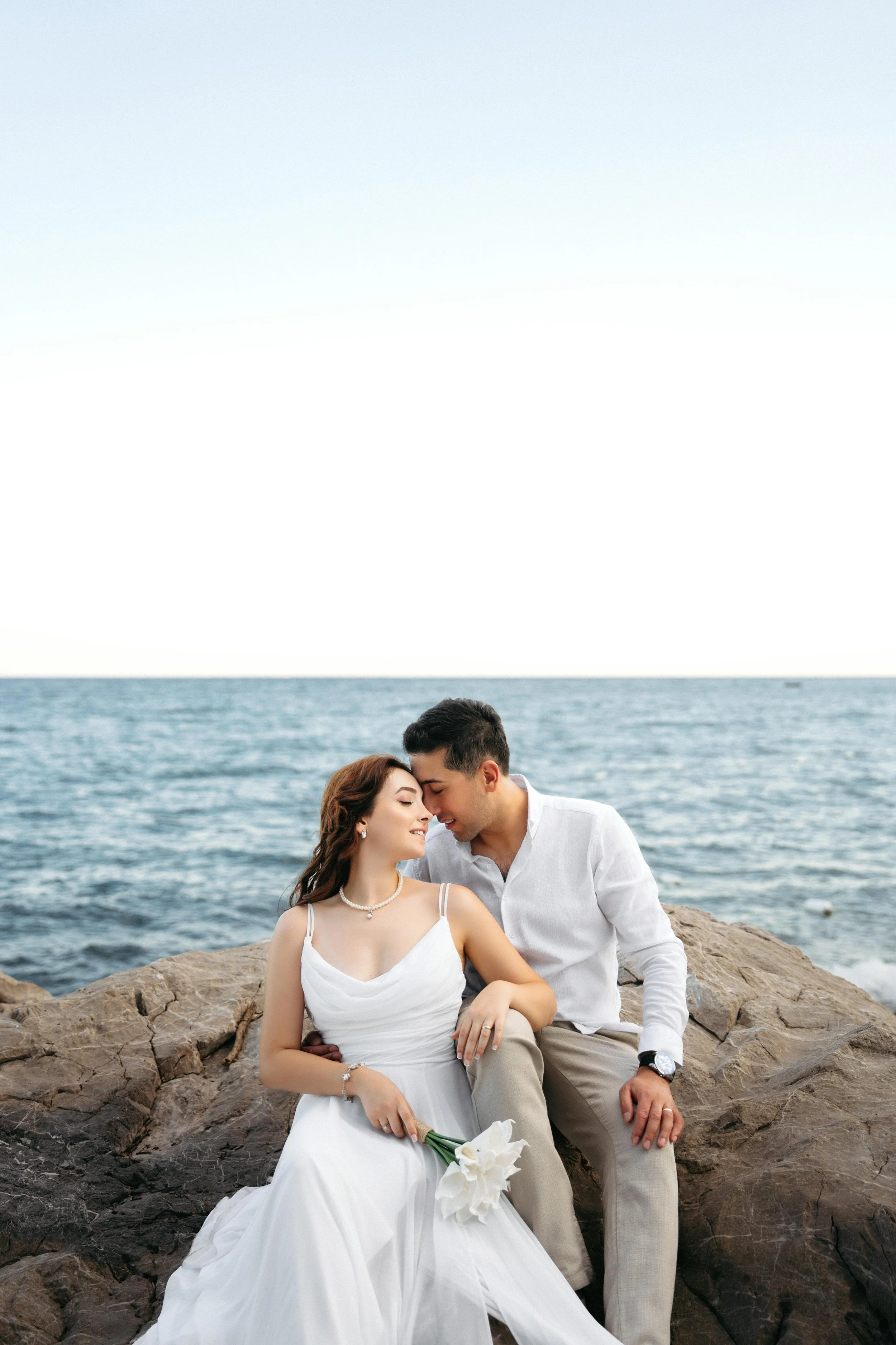A couple dressed in wedding attire sitting on rocks by the ocean, smiling and leaning close together with foreheads touching.