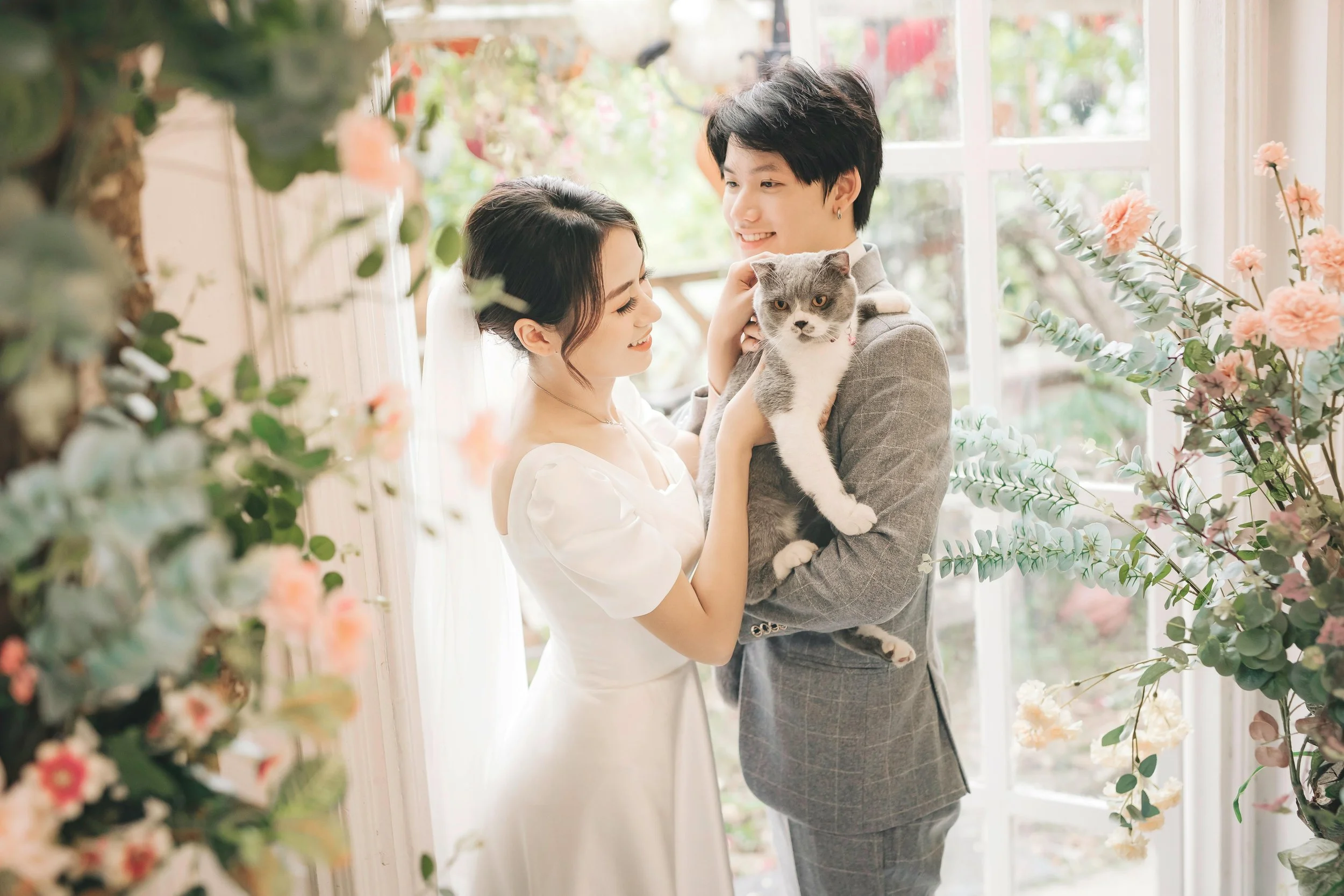 A bride and groom holding a cat inside a bright, floral decorated room with large windows.