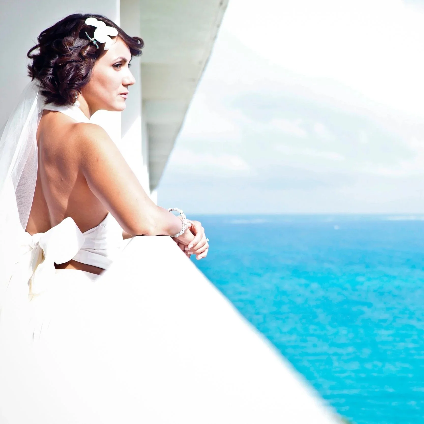 A woman with dark, styled hair and a white flower hair accessory, wearing a white dress with a backless design, leaning on a balcony railing, looking at the ocean view.