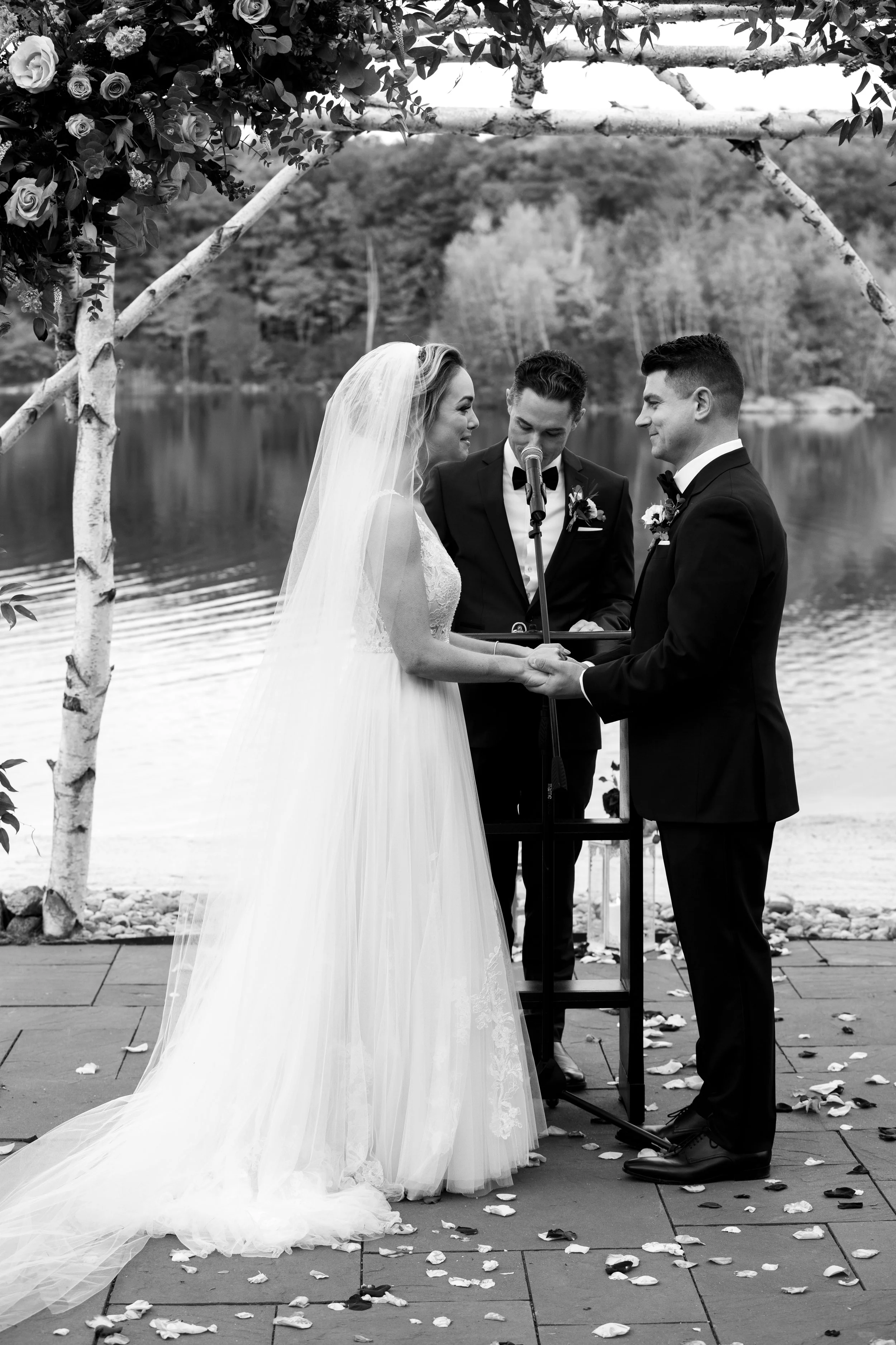 A black and white photo of a wedding ceremony by a lake, with a bride and groom holding hands and facing each other, an officiant standing between them, and an arch decorated with flowers and greenery above.
