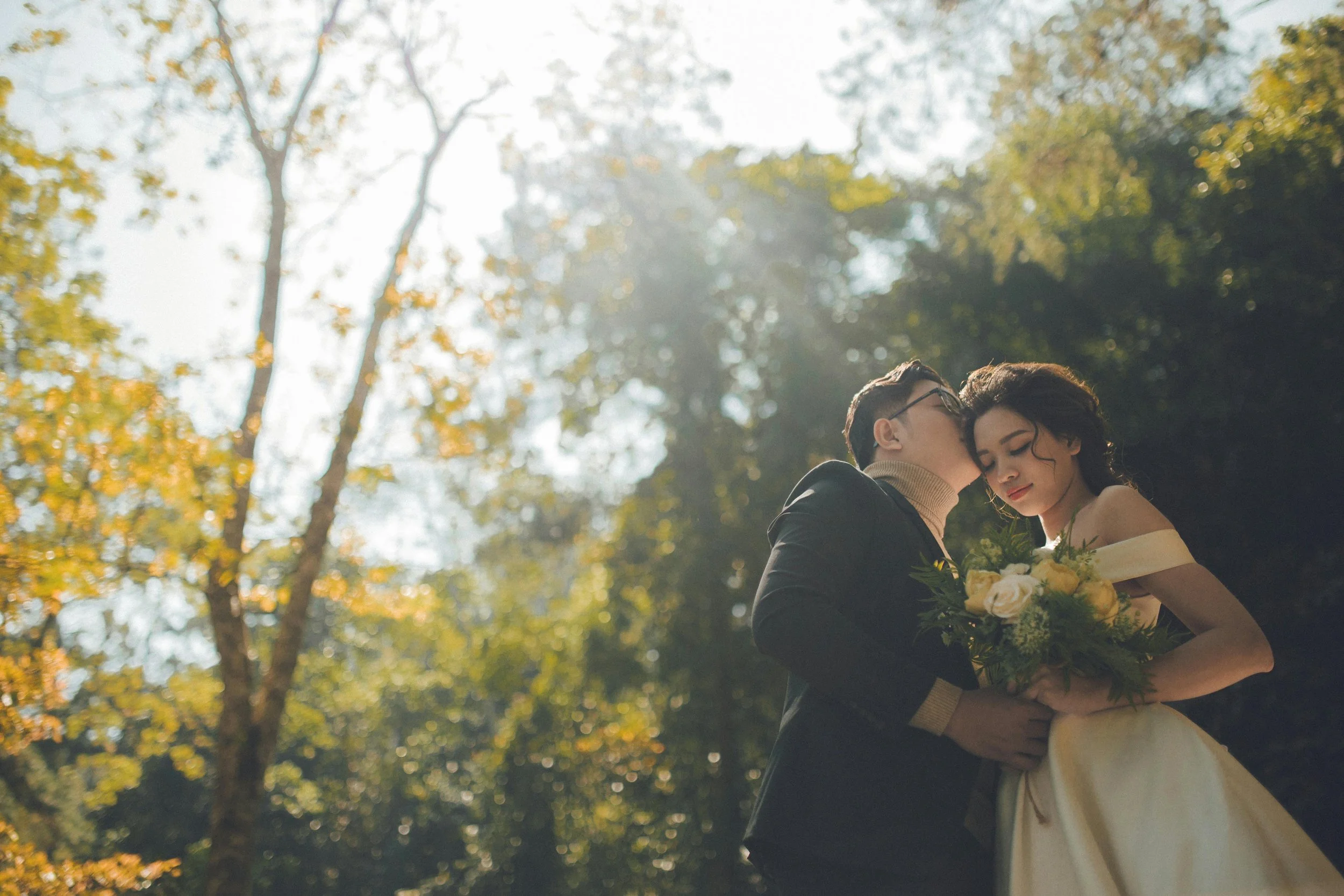 A couple in wedding attire sharing a tender moment outdoors, the man gently kissing the woman's forehead, with trees and sunlight creating a dreamy background.