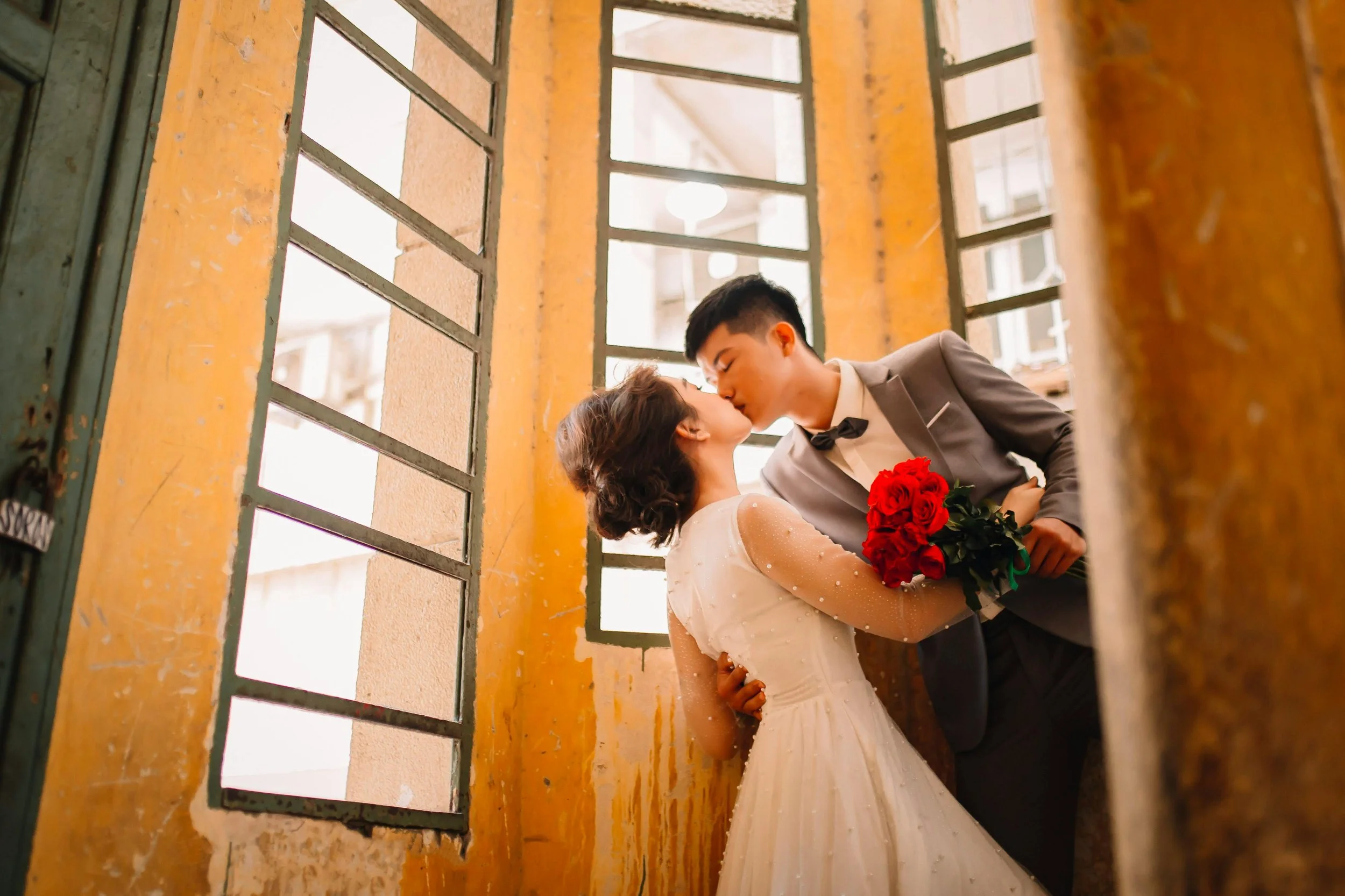 A bride and groom share a kiss in a rustic setting, with the groom holding a bouquet of red roses. The photo is taken through yellow window bars.